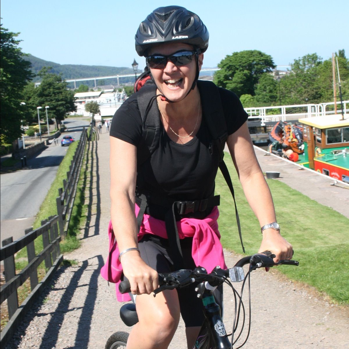 Woman in helmet and sunglasses rides a scooter on a path beside a canal.