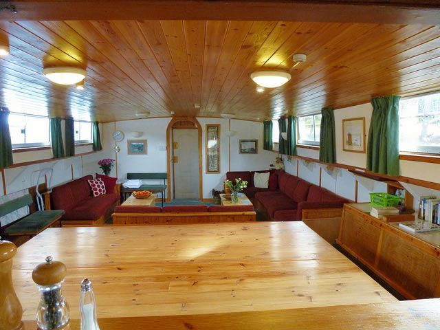 Interior of a boat with wooden walls and ceiling. Red sofas line the walls, a kitchen area visible.