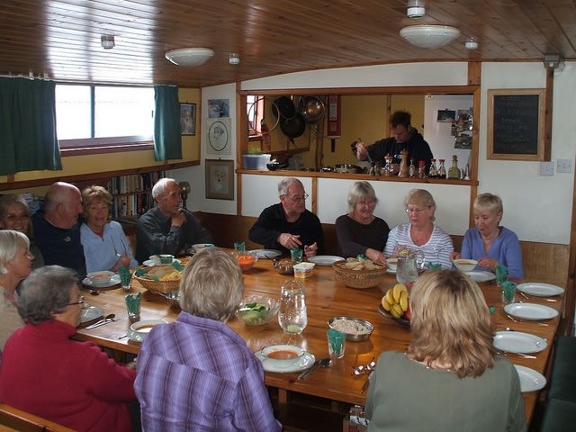 People seated around a large wooden table, eating in a room with a kitchen area and window.