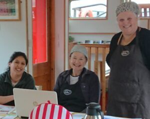 Three people smiling at a counter with a laptop, a red and white striped tea cozy, and a coffee pot in a room.