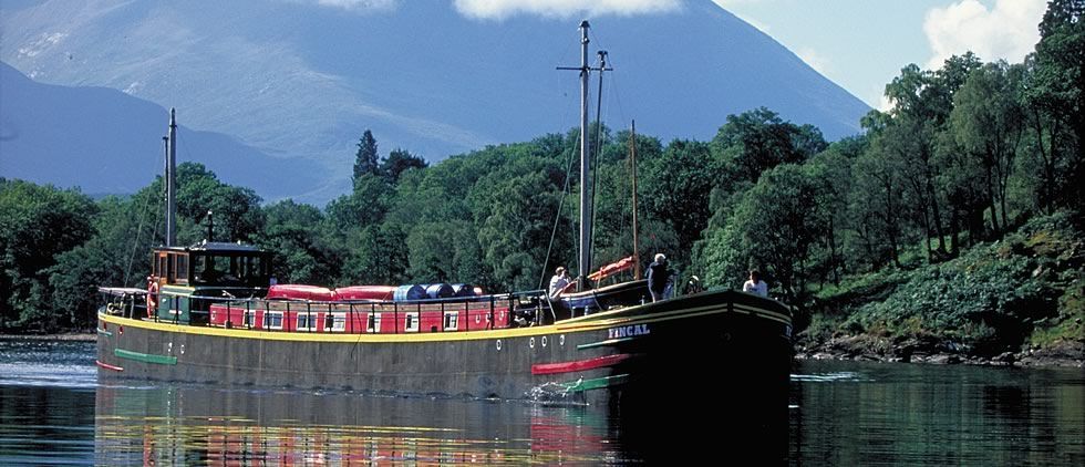 A long boat on water with red structures, surrounded by green trees and mountains in the background.