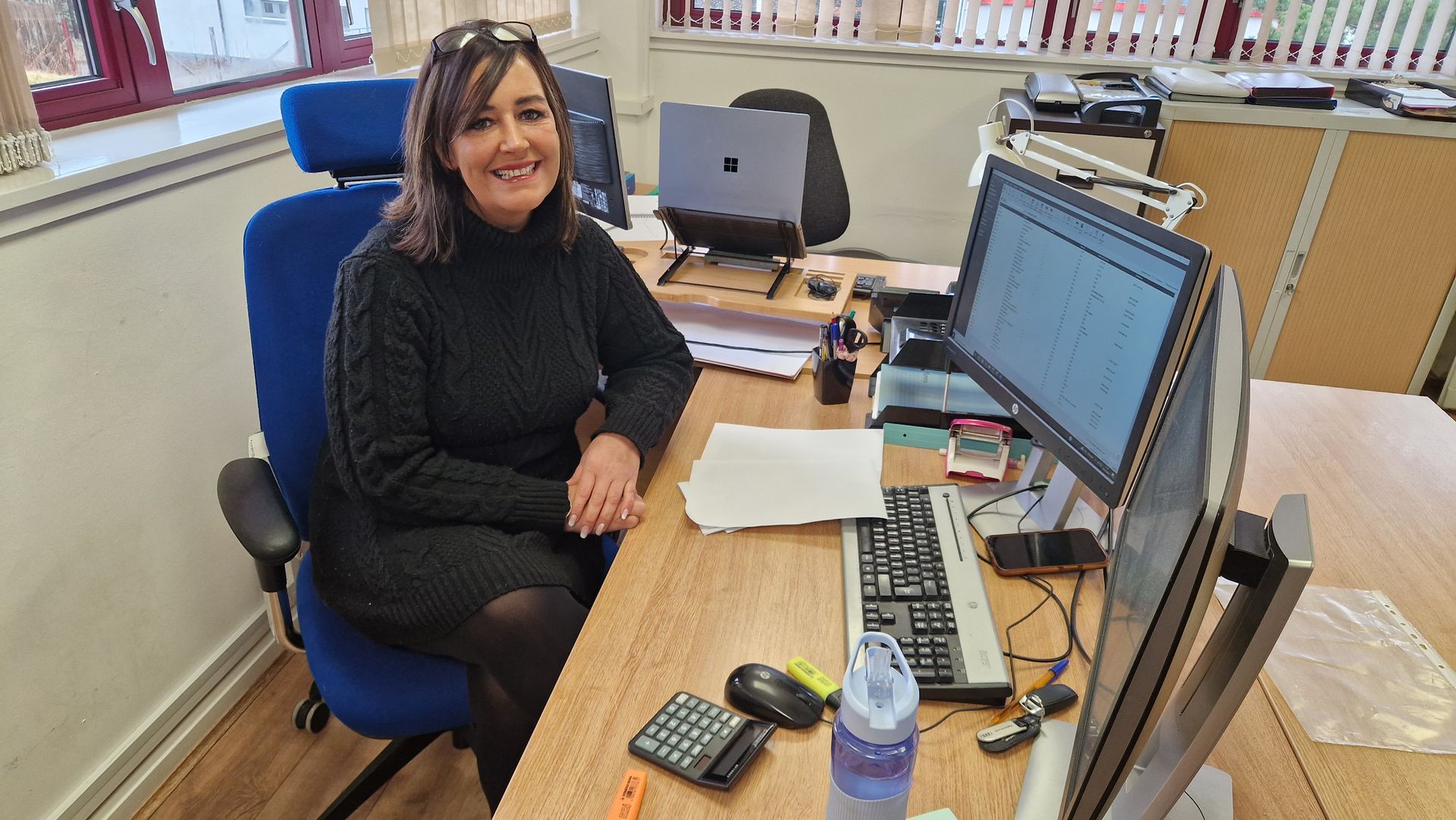 Woman seated at desk, smiling, in an office. Laptop, computer monitor, keyboard, and other office supplies present.
