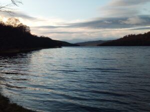 Calm lake scene at dusk, framed by dark forested shores under a cloudy sky.