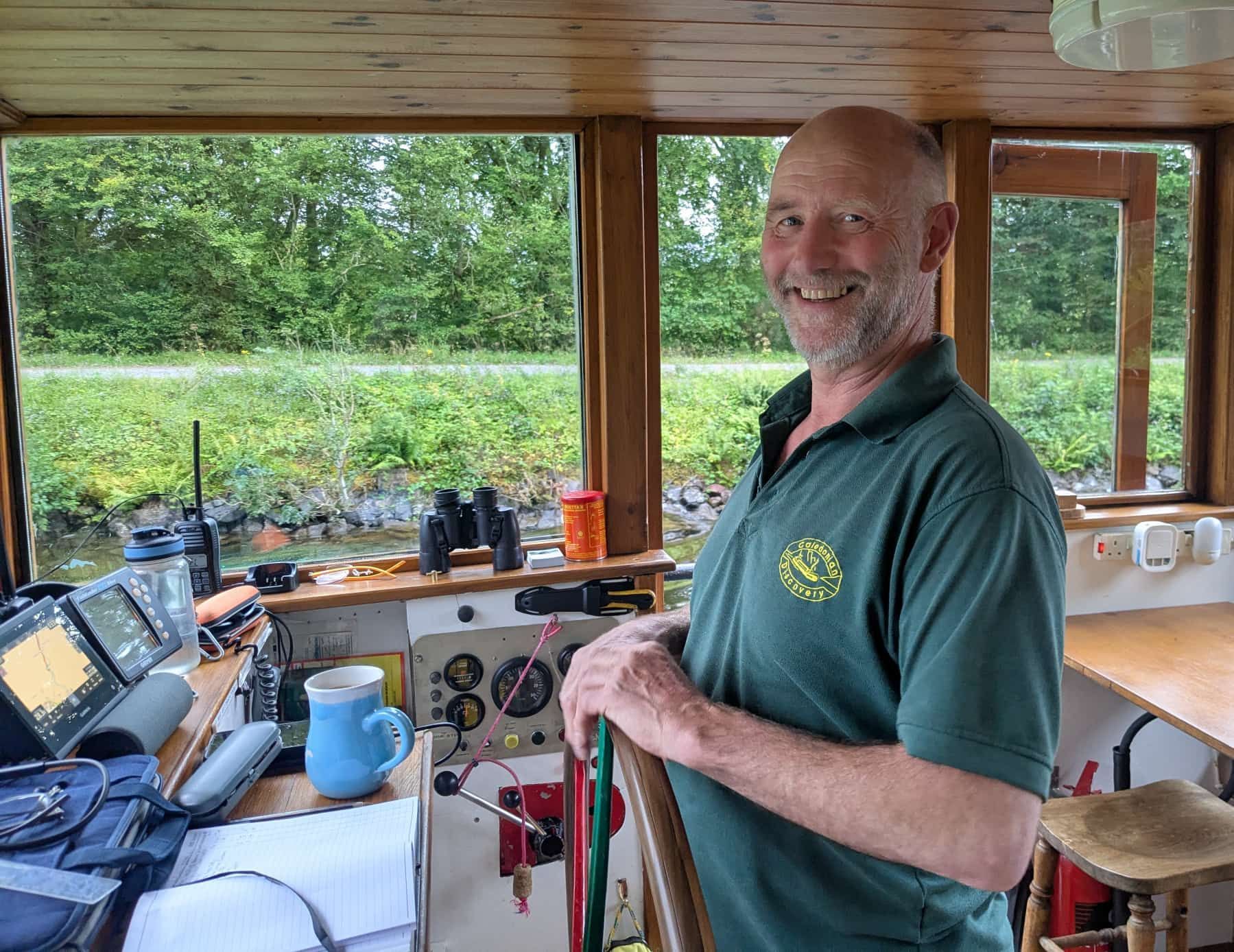 Man smiling at the wheel of a boat, in a wooden cabin. He wears a green shirt.