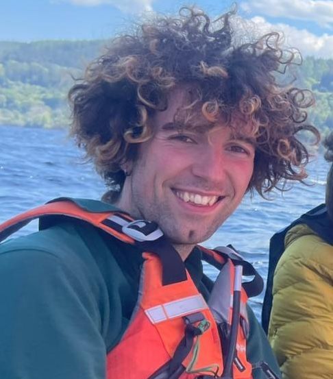 Man with curly hair smiling, wearing orange life vest, on a boat, against lake and mountains.