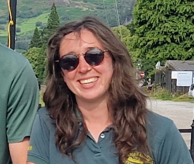 Woman with sunglasses smiles outdoors, wearing a dark green shirt.