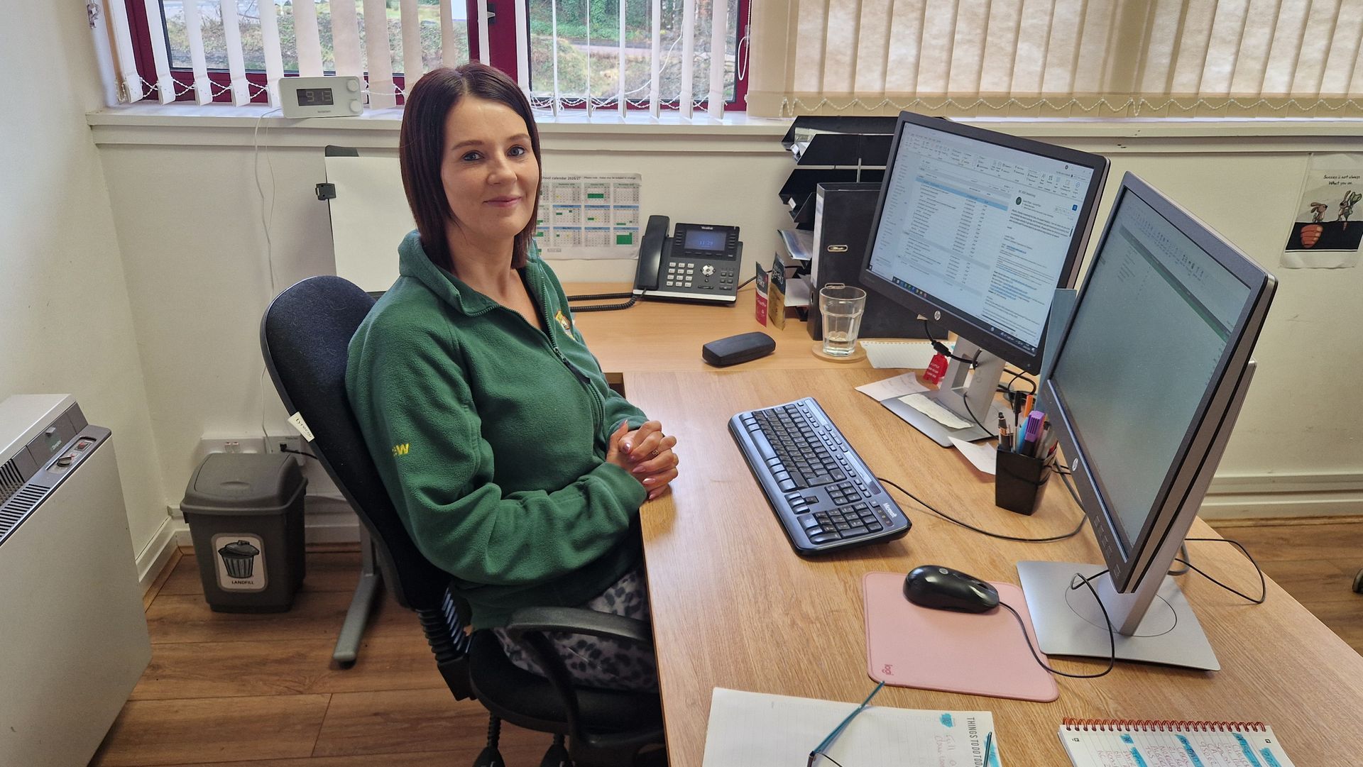 Woman seated at desk with two computer monitors, smiling. Office setting.