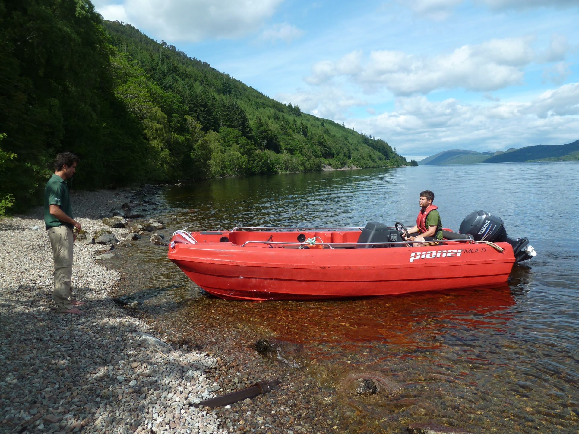 A red motorboat with two men on a rocky shore, near a forested hillside on a lake.