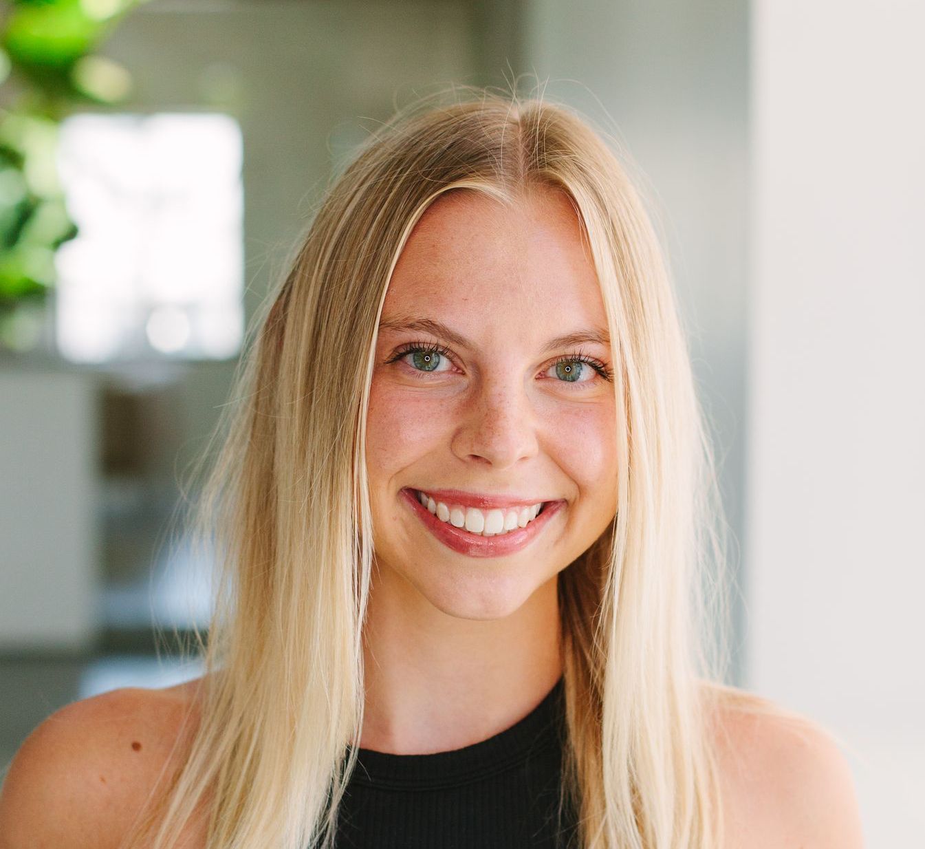 Young woman with long blonde hair, smiling at the camera in a bright, indoor setting. She wears a black top.