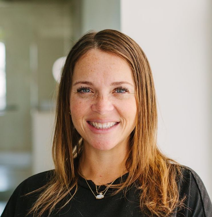 Smiling woman with light brown hair, wearing a black shirt and necklace, in a well-lit room.
