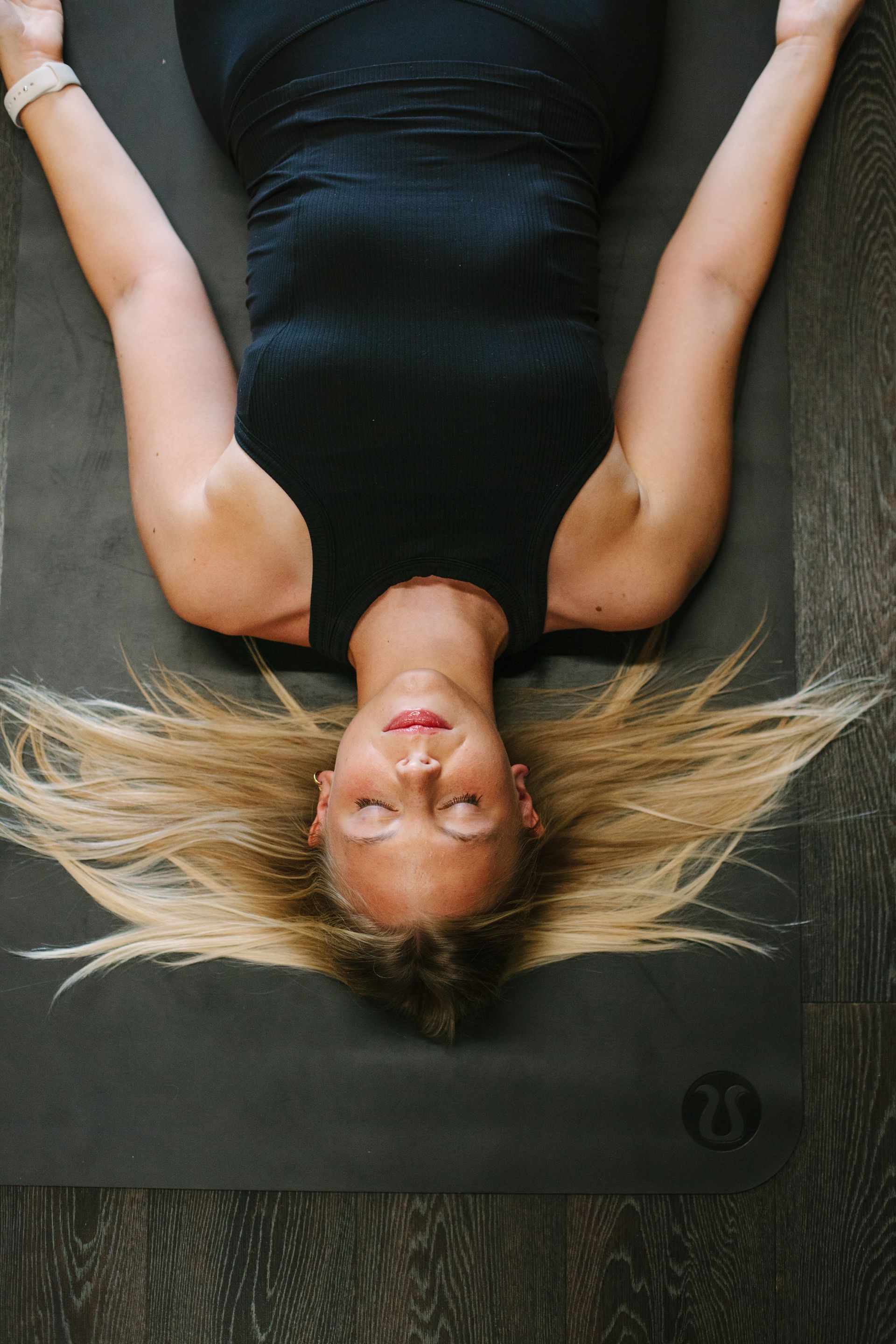 Woman in yoga pose lying on a mat with arms out, blonde hair, black top.