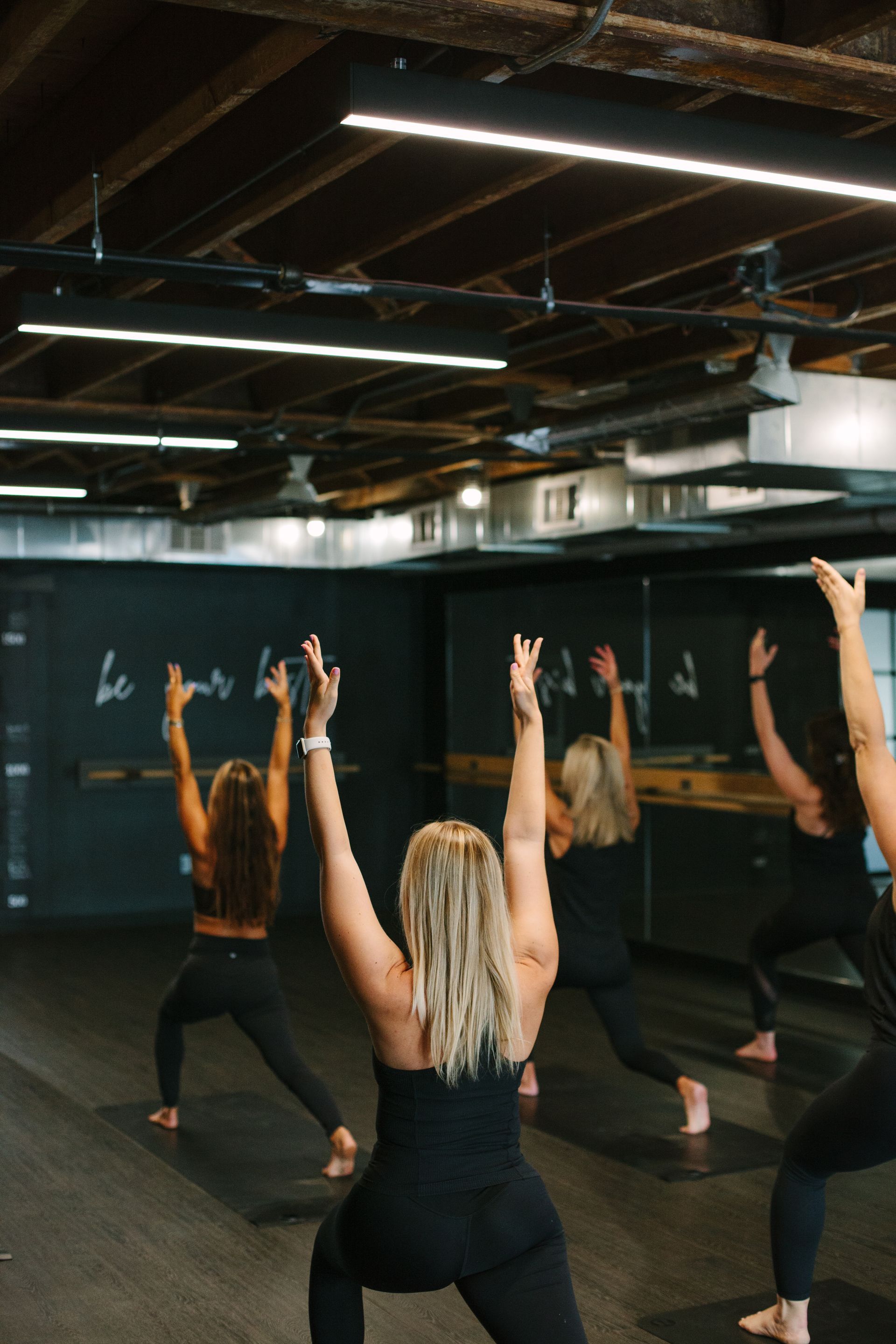 Women in a yoga class stretch, raising arms in a dim studio.