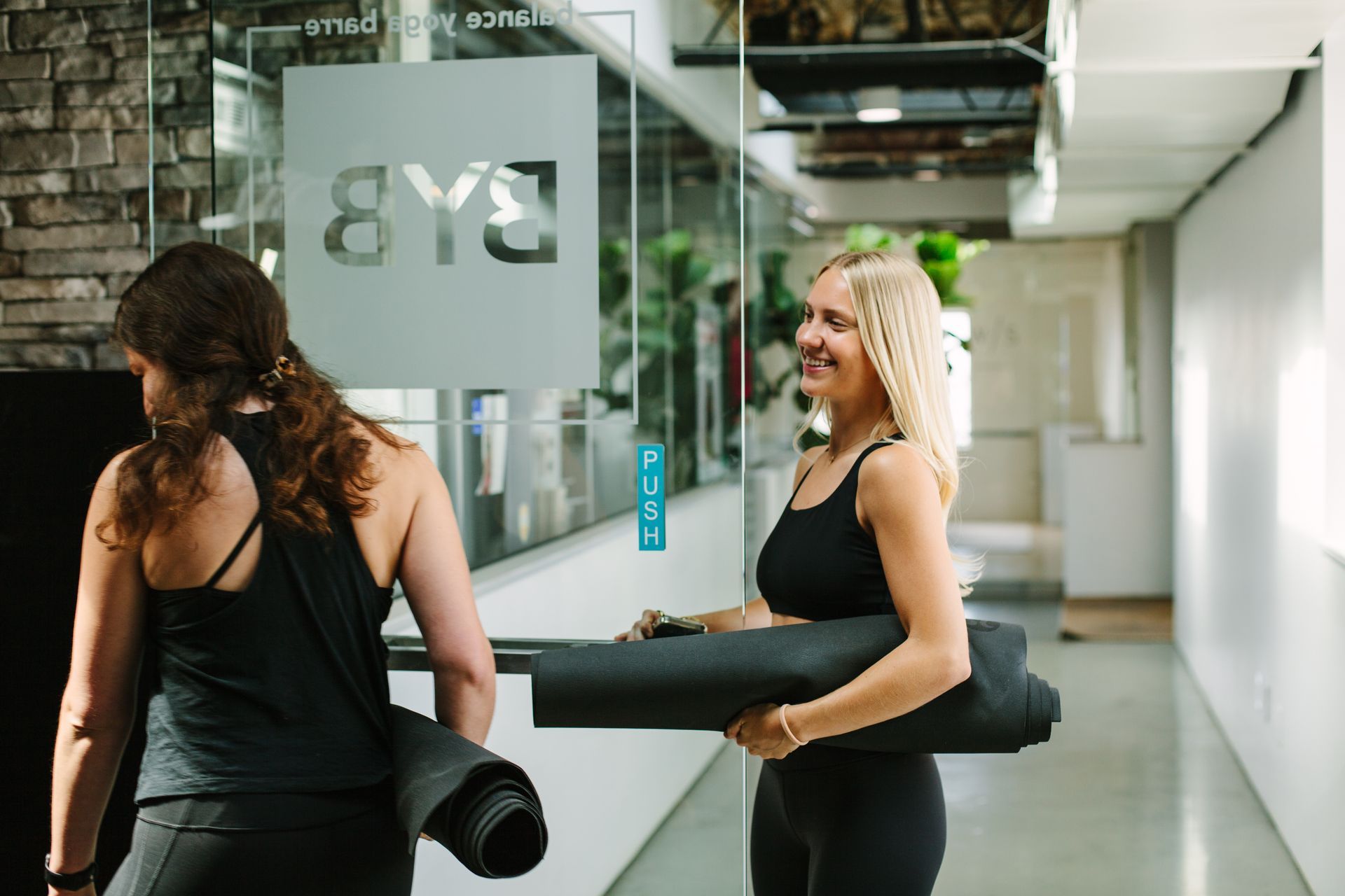 Two women, carrying yoga mats, enter a studio with