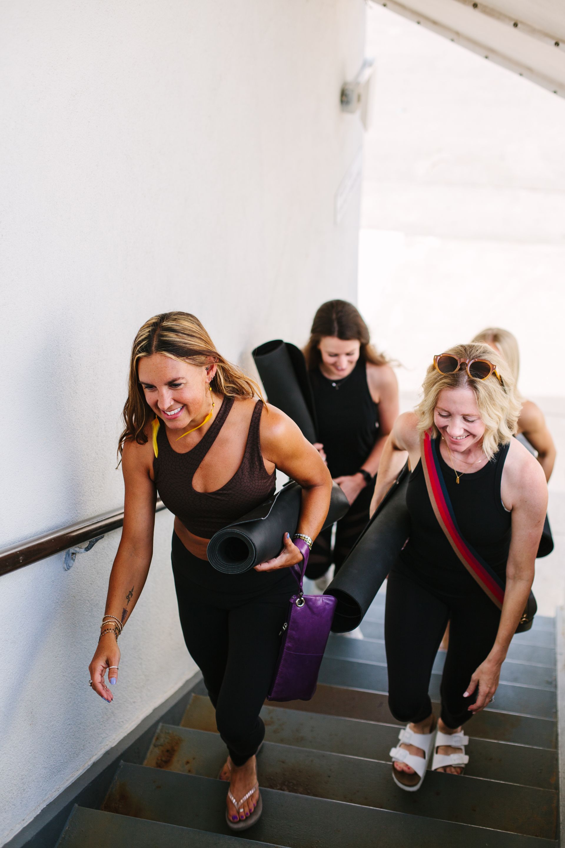 Four women ascend stairs, carrying yoga mats. Smiling, wearing workout clothes in a white setting.