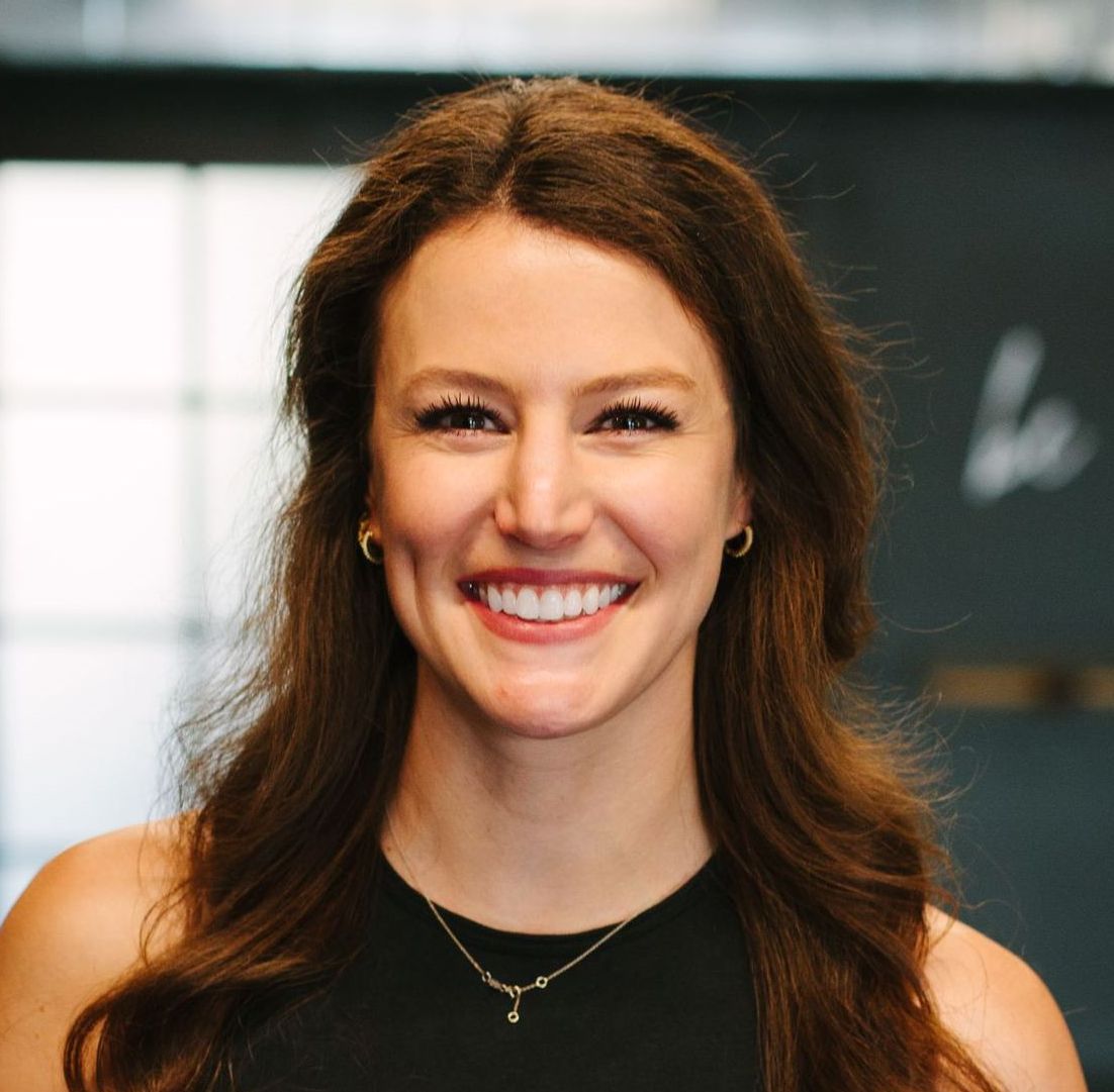 Woman with long brown hair, smiling at the camera, wearing a black top and a necklace.