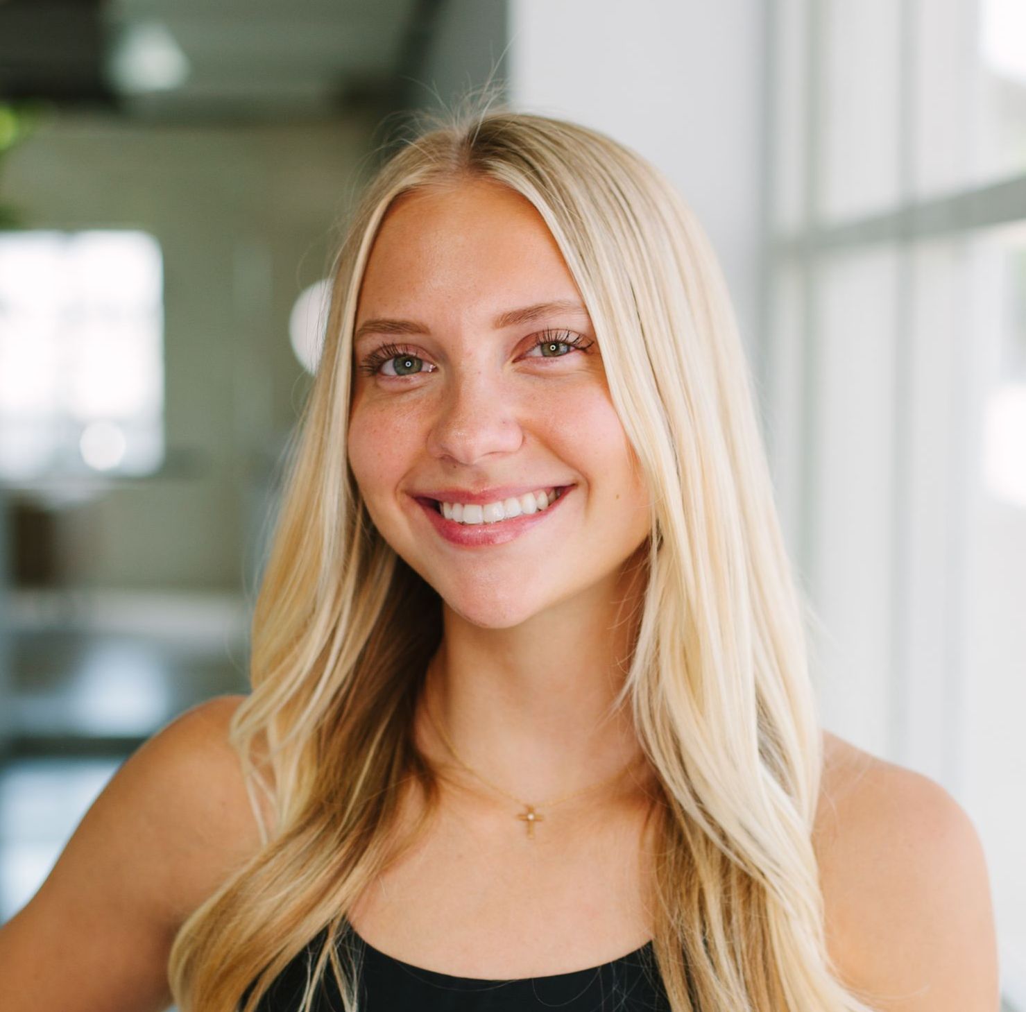 Blonde woman smiling at the camera, wearing a black tank top and a necklace. Indoors with natural light.