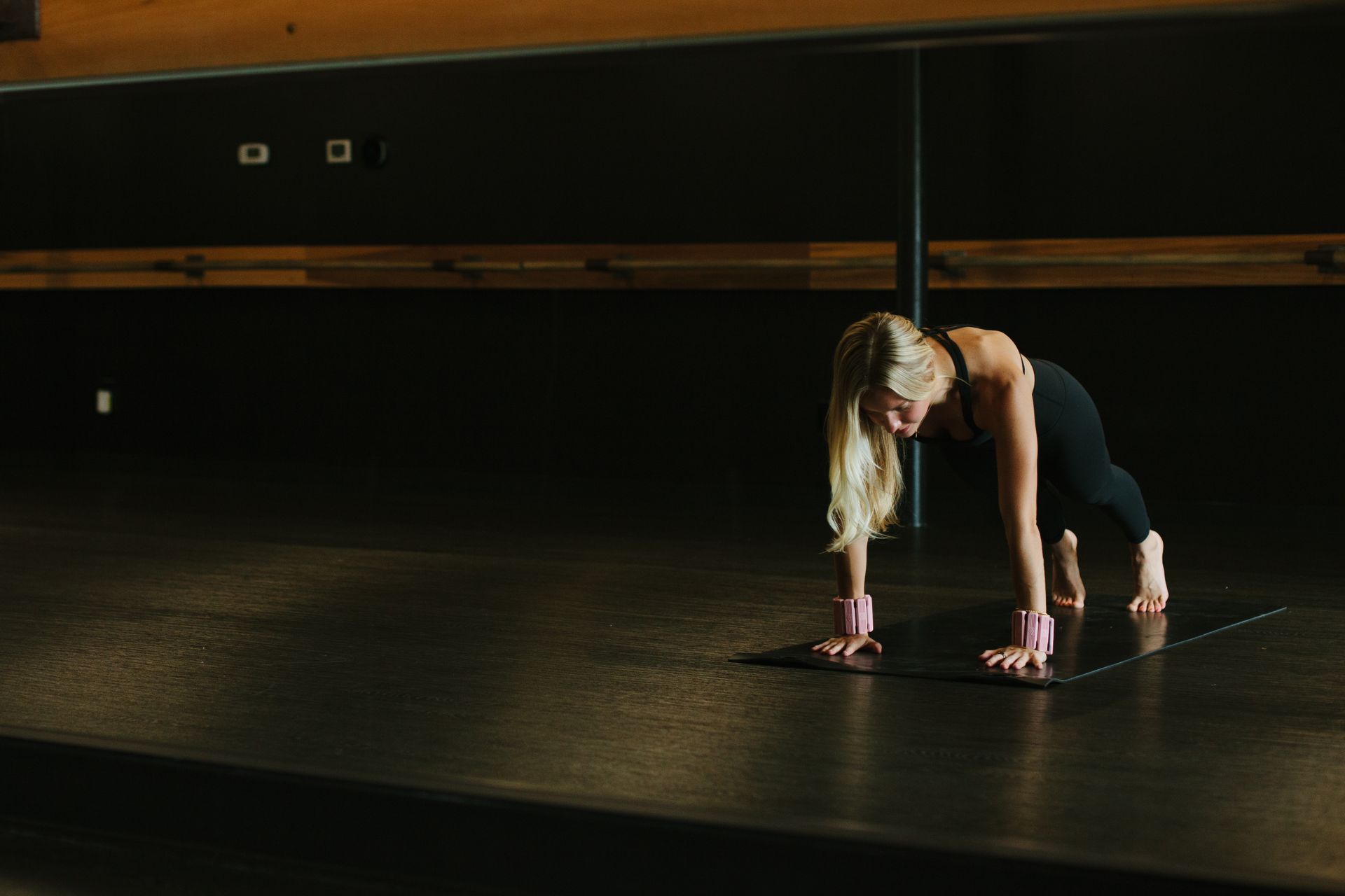 Blonde woman in a plank pose on a yoga mat in a dark studio; ankle weights.
