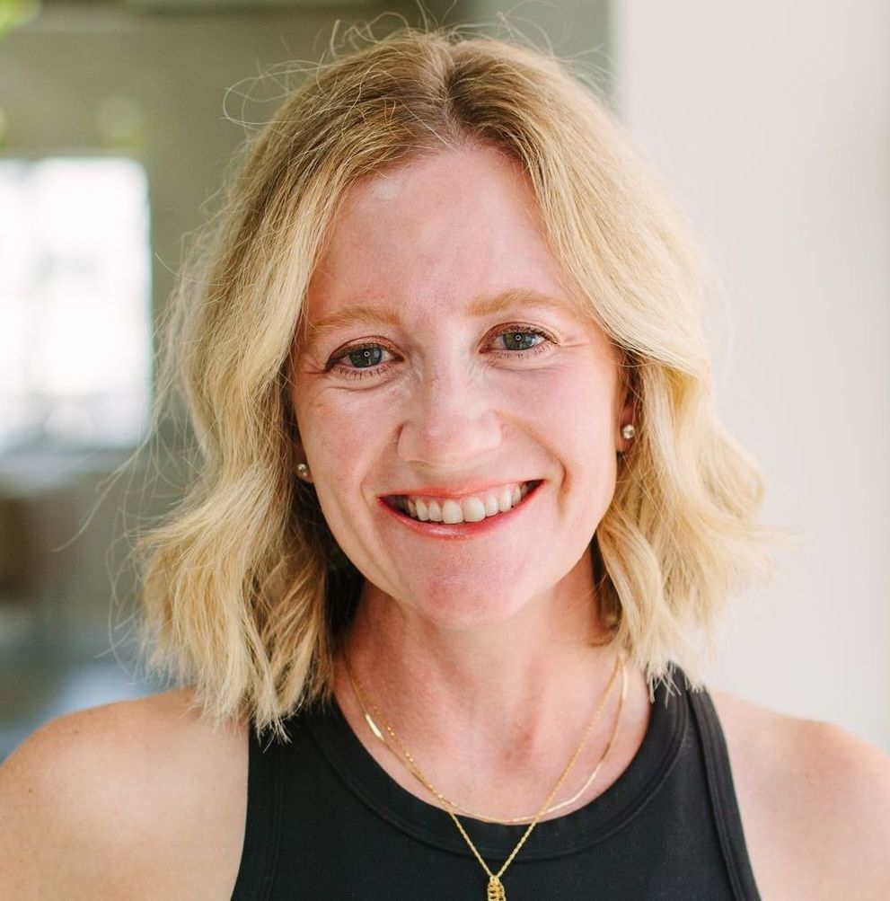 Blonde woman smiling, wearing a black top and gold necklace, outdoors.