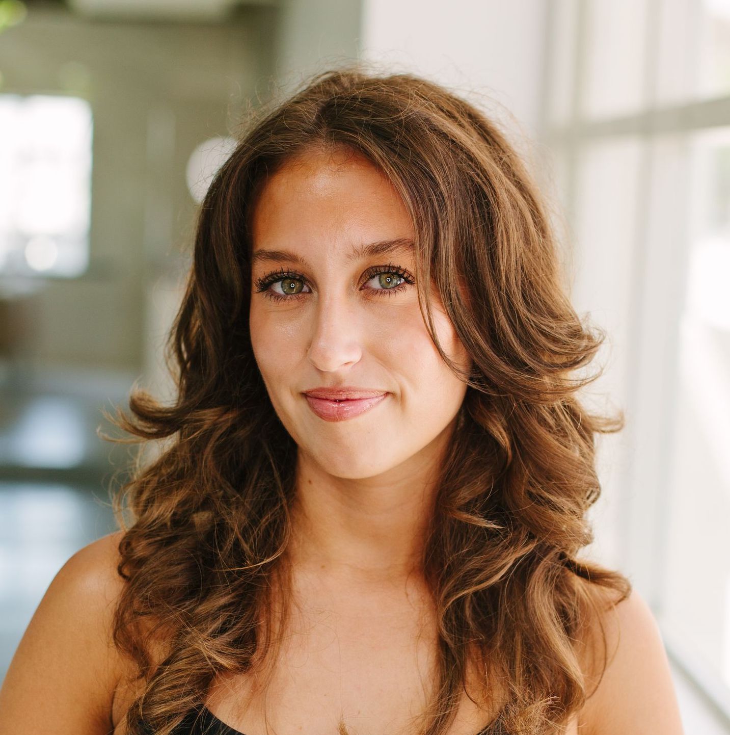 Woman with curly brown hair smiles slightly at the camera. She is in a bright room.