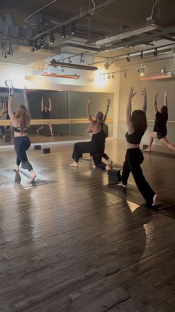 A group of people in a yoga studio performing high lunges with arms raised, mirrored in the background.