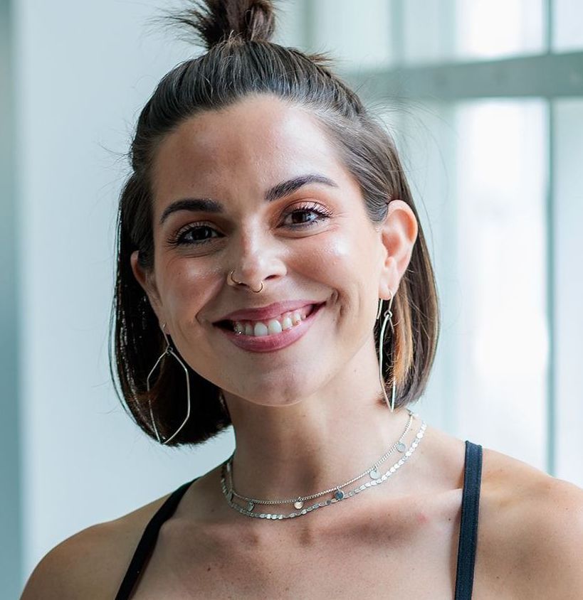 Smiling woman with a nose ring and a high bun, wearing dangling earrings and two layered necklaces, in a well-lit room.