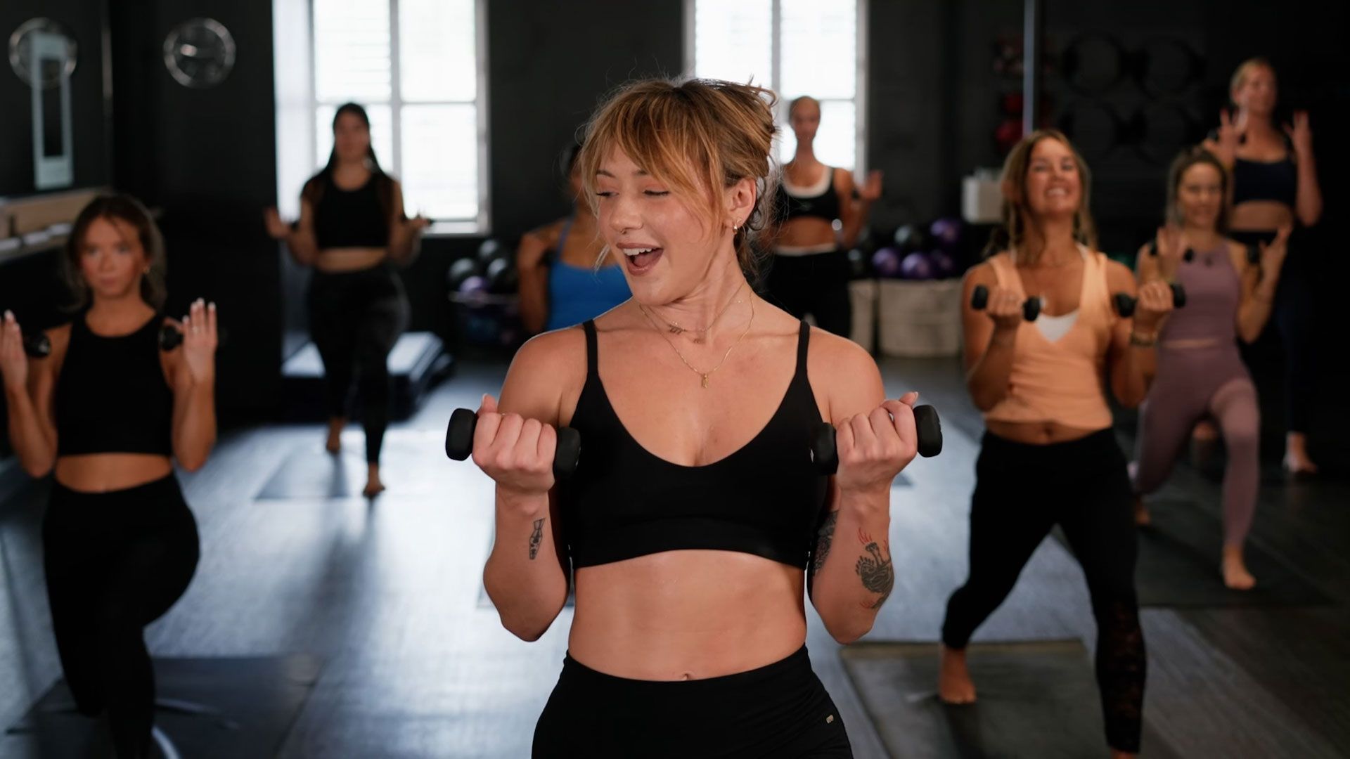 A woman leads a workout class, holding dumbbells. Others follow along, lifting weights in a studio.