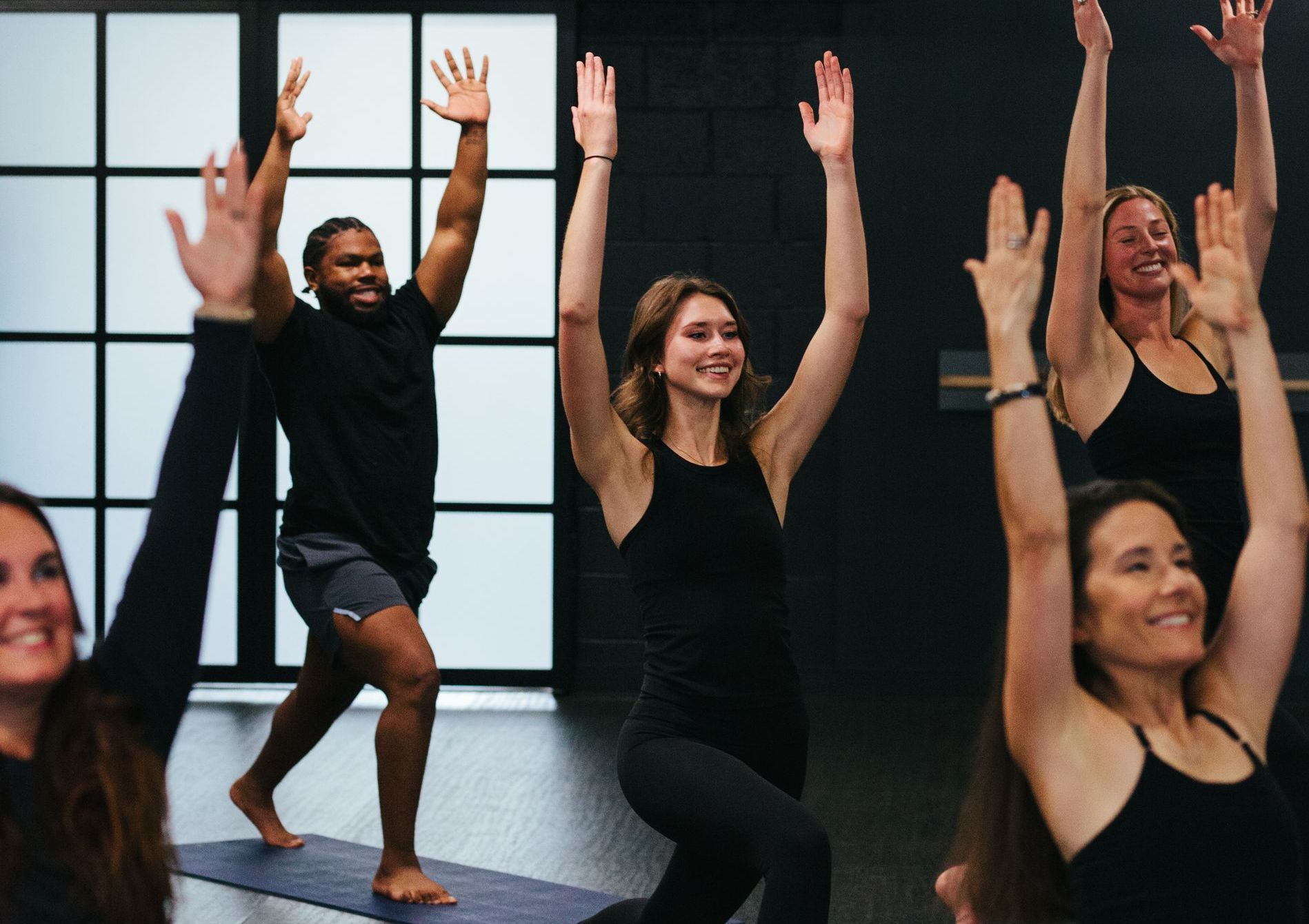 People in a yoga class raising arms in a lunge, smiling. Black clothing, studio with windows.