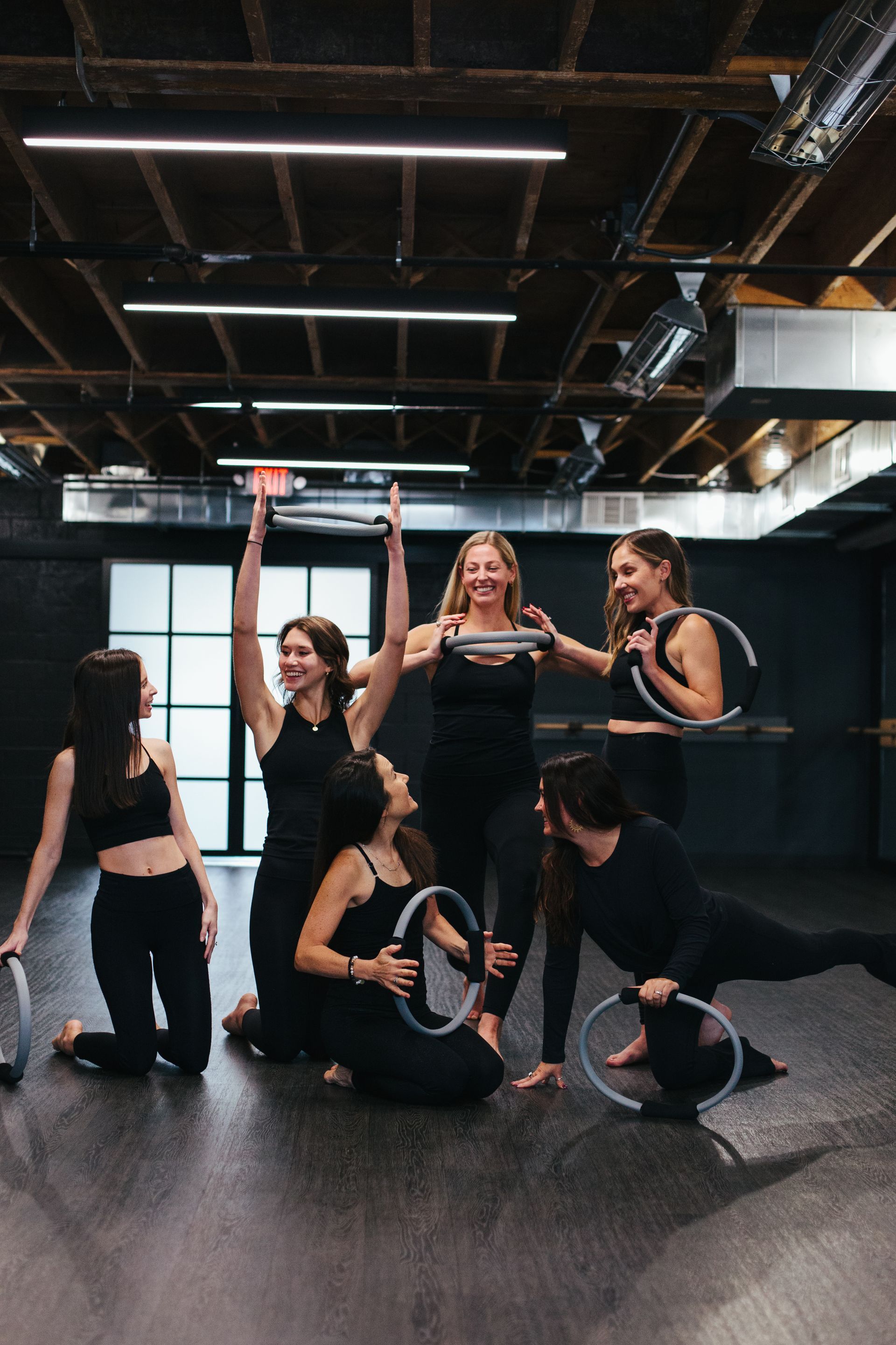Group of women in black workout attire pose with Pilates rings in a studio. Some hold rings overhead or at waist level.