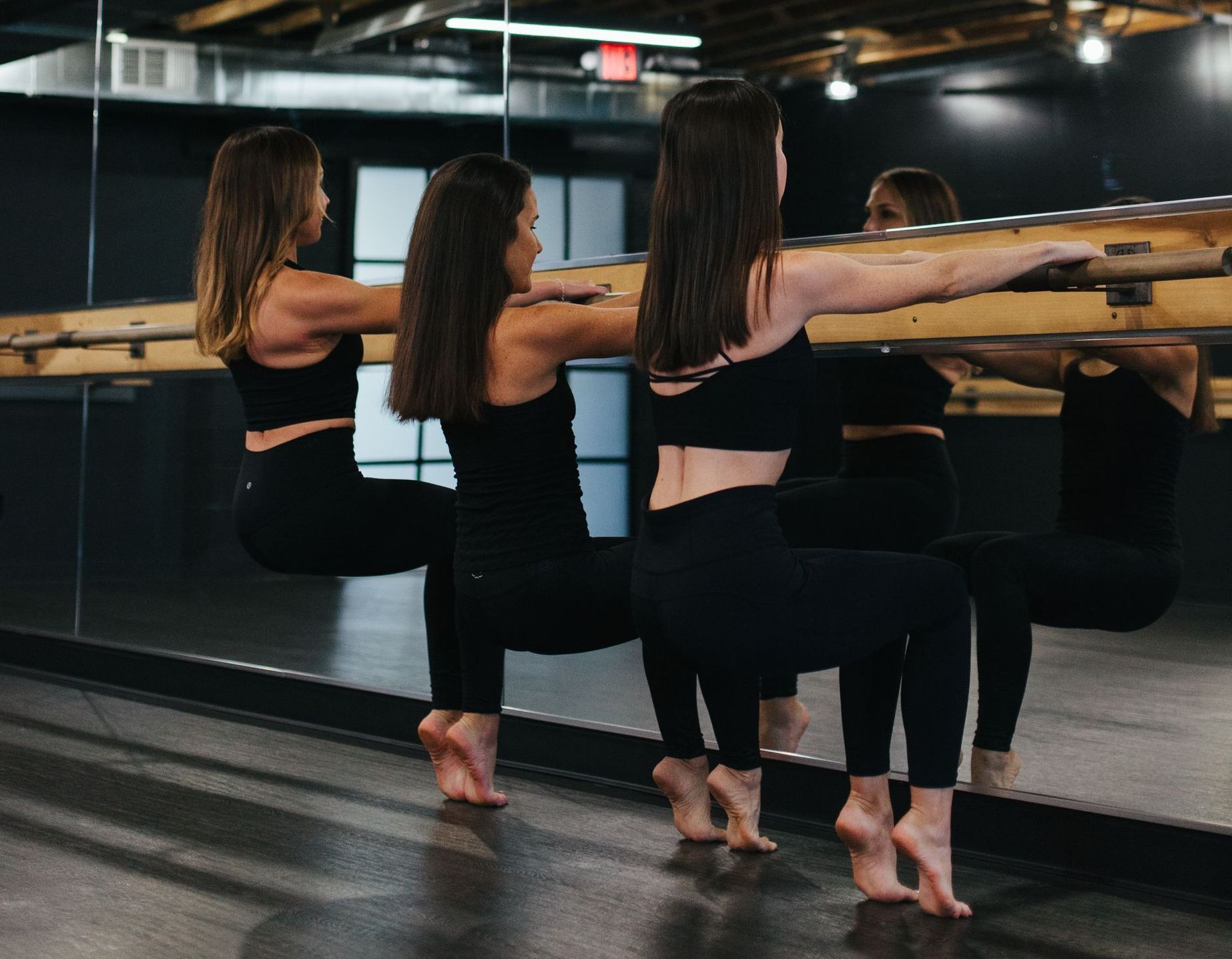 Three women in black activewear doing a barre exercise, holding onto a wooden bar in a studio with a mirrored wall.