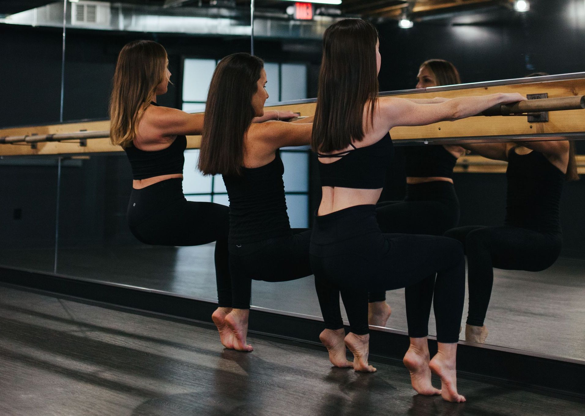 Three women in black workout clothes doing a barre exercise, holding a barre, in a studio with a mirrored wall.