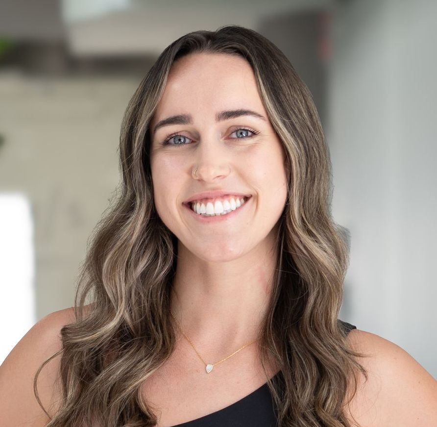 Woman with wavy brown hair smiles at the camera, wearing a black top and a gold necklace.