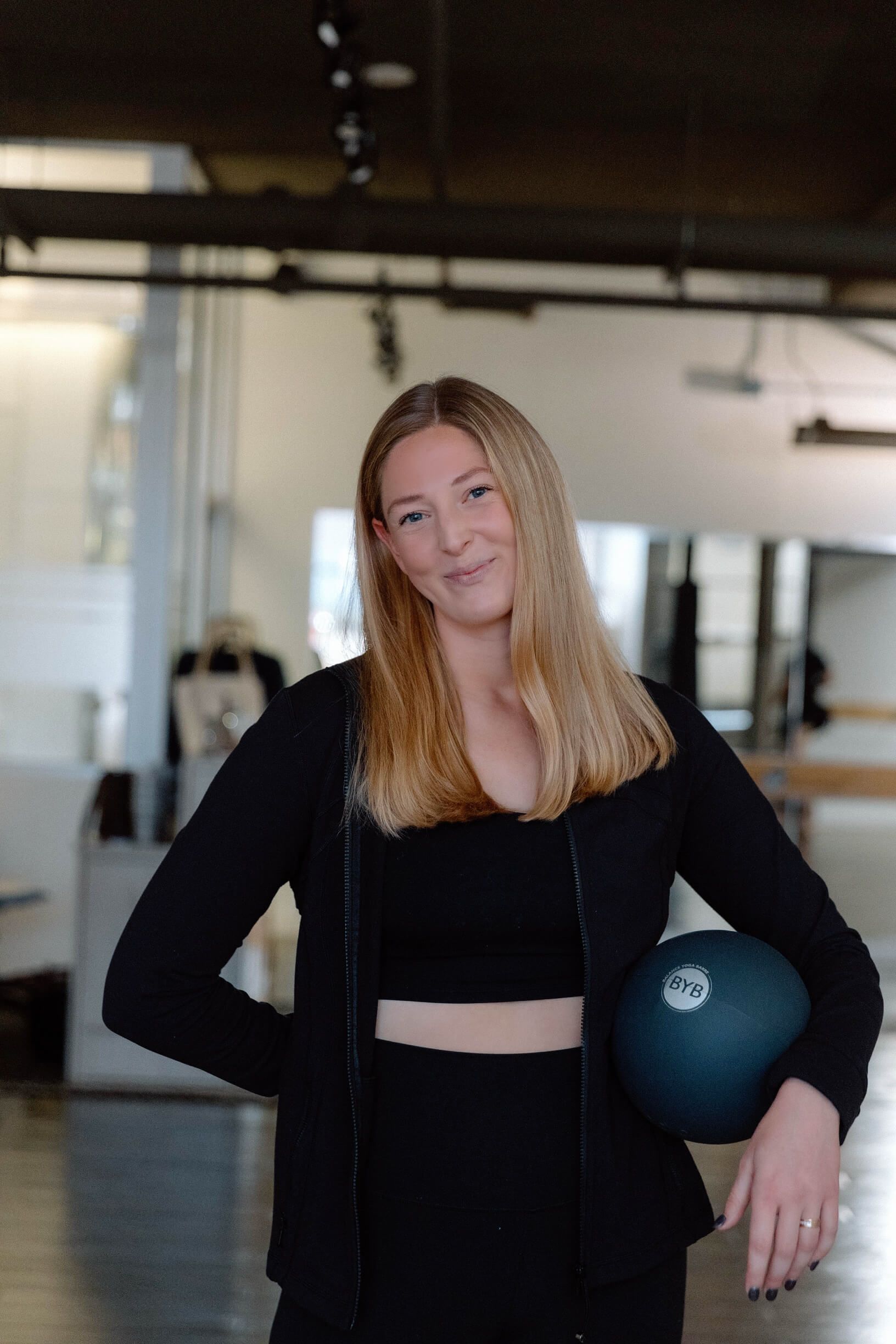 A smiling person with long blonde hair, wearing a black outfit, holds a dark blue exercise ball in a gym setting.