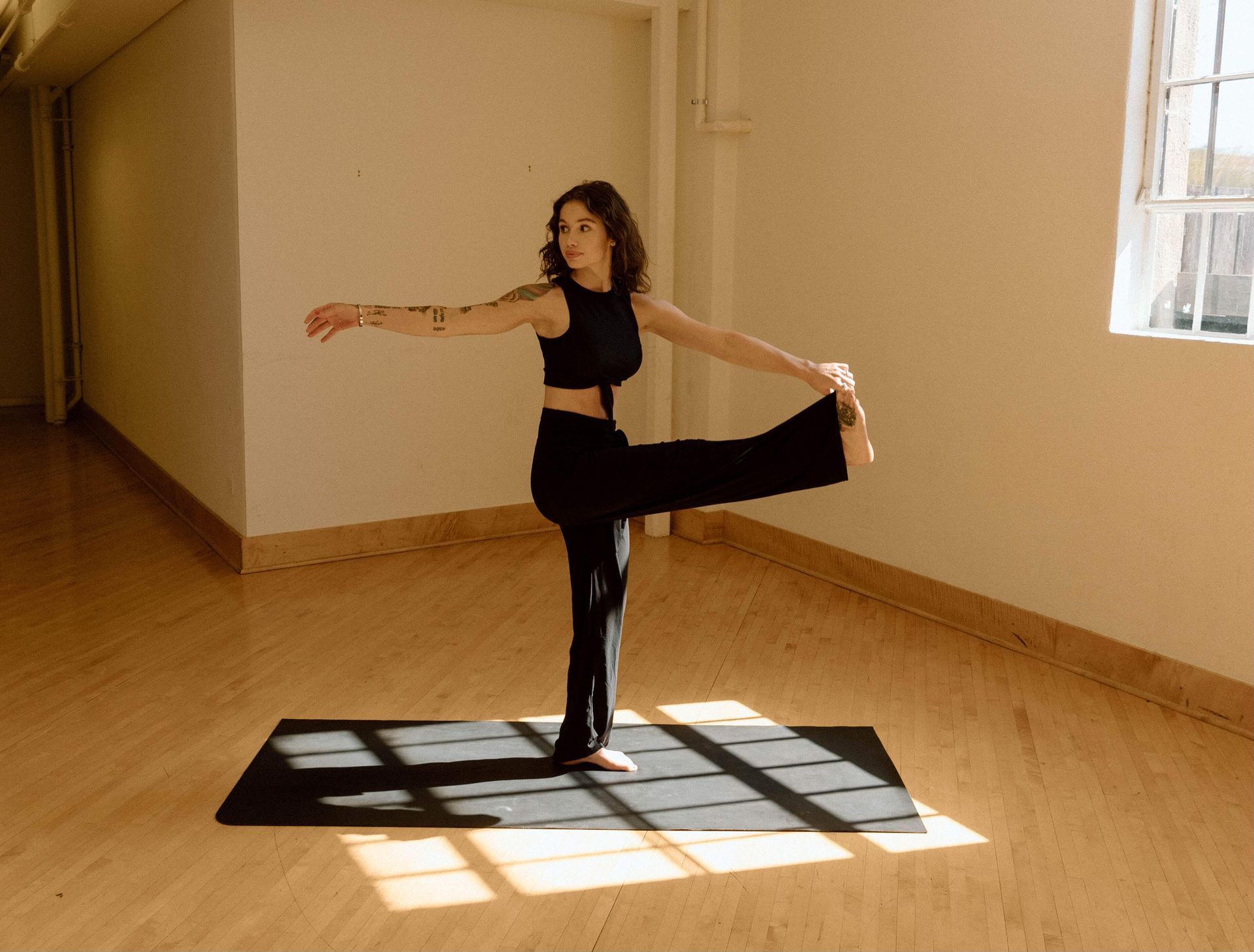 A person in a black yoga outfit performs a standing hand-to-big-toe pose on a mat in a bright, minimalist studio.