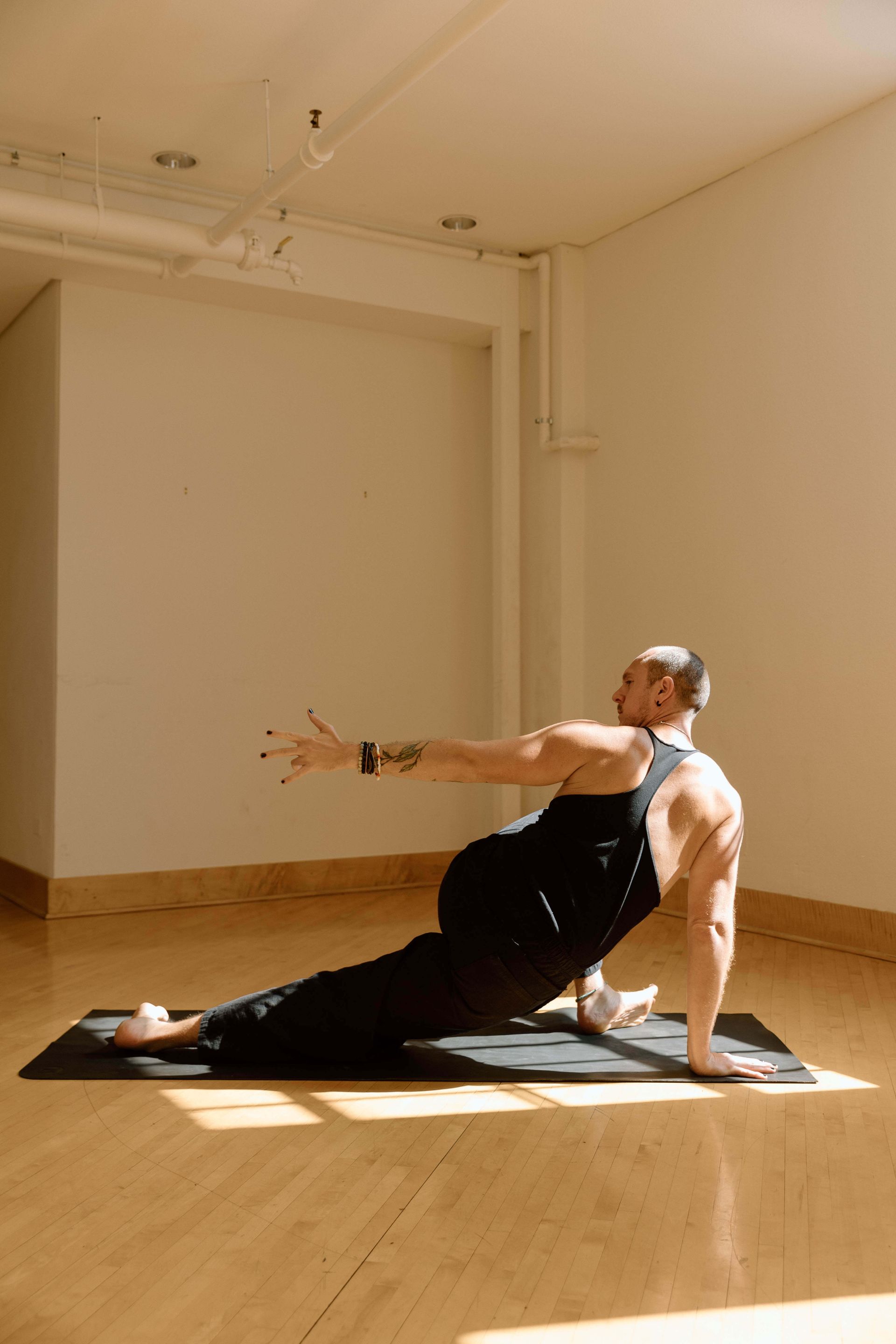 A person in black workout gear on a yoga mat, performing a low lunge with a torso twist and reaching arm in a bright studio.