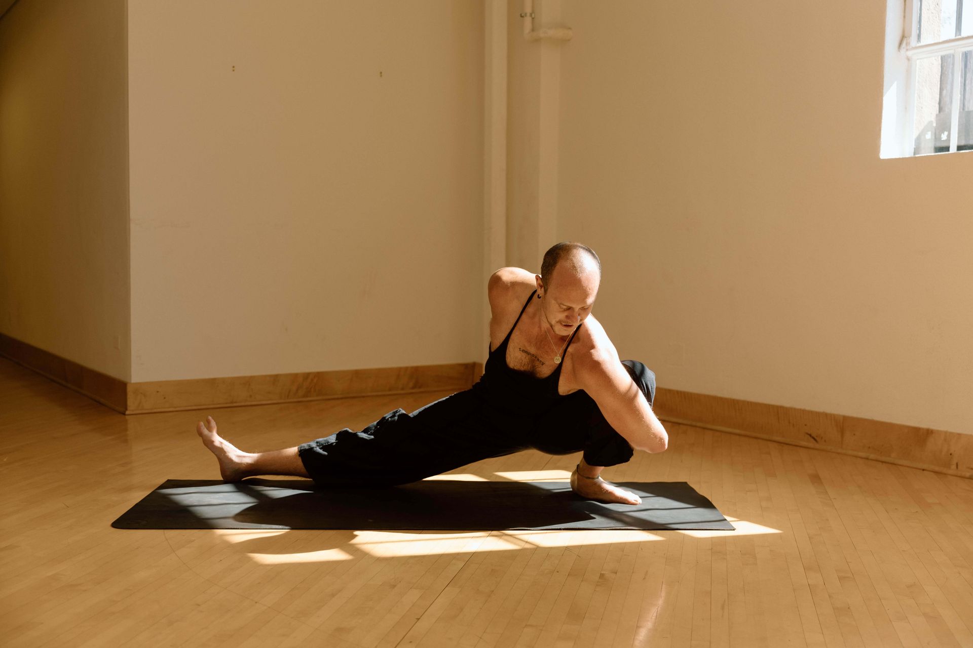 A person in black clothing performs a deep side lunge yoga pose with a torso twist on a mat in a bright room.