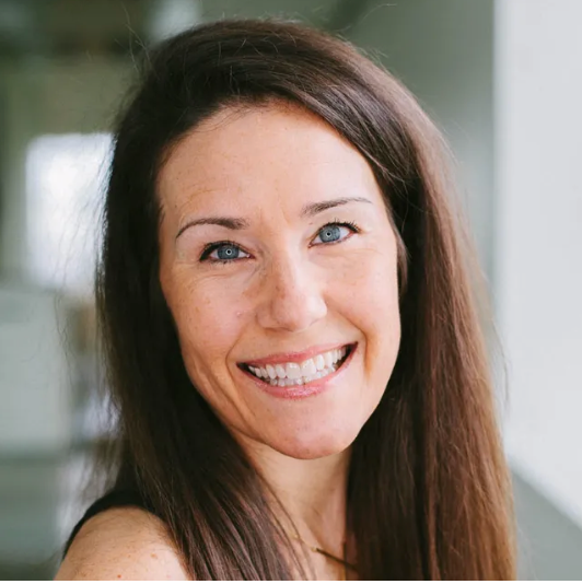 Woman with long brown hair smiles at the camera, blue eyes, neutral background.