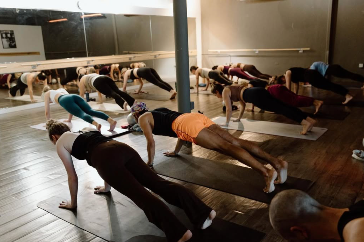 People in a yoga class doing plank pose on mats in a studio with a mirror.