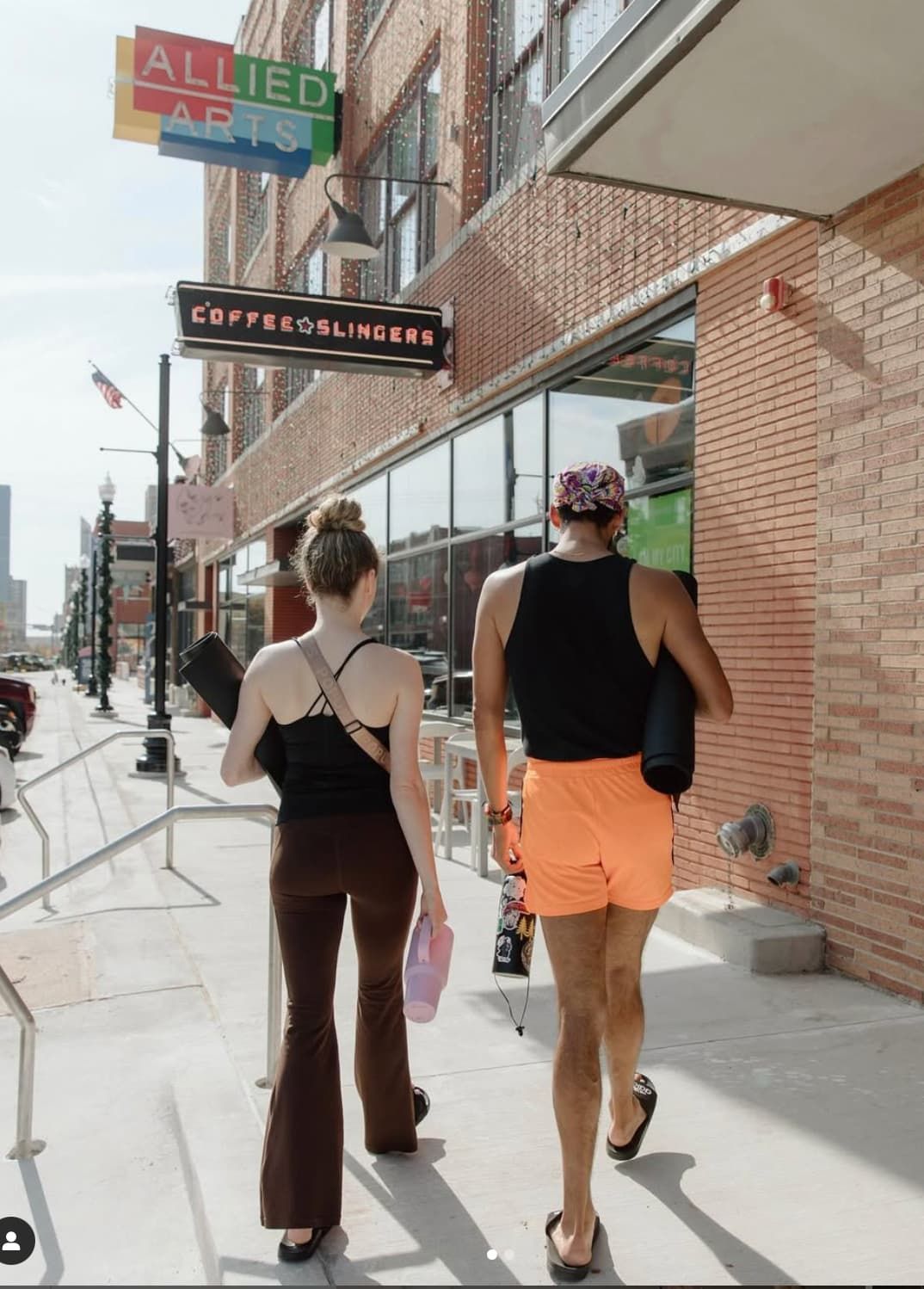 Two people with yoga mats walk down a sunny city sidewalk past a brick building with