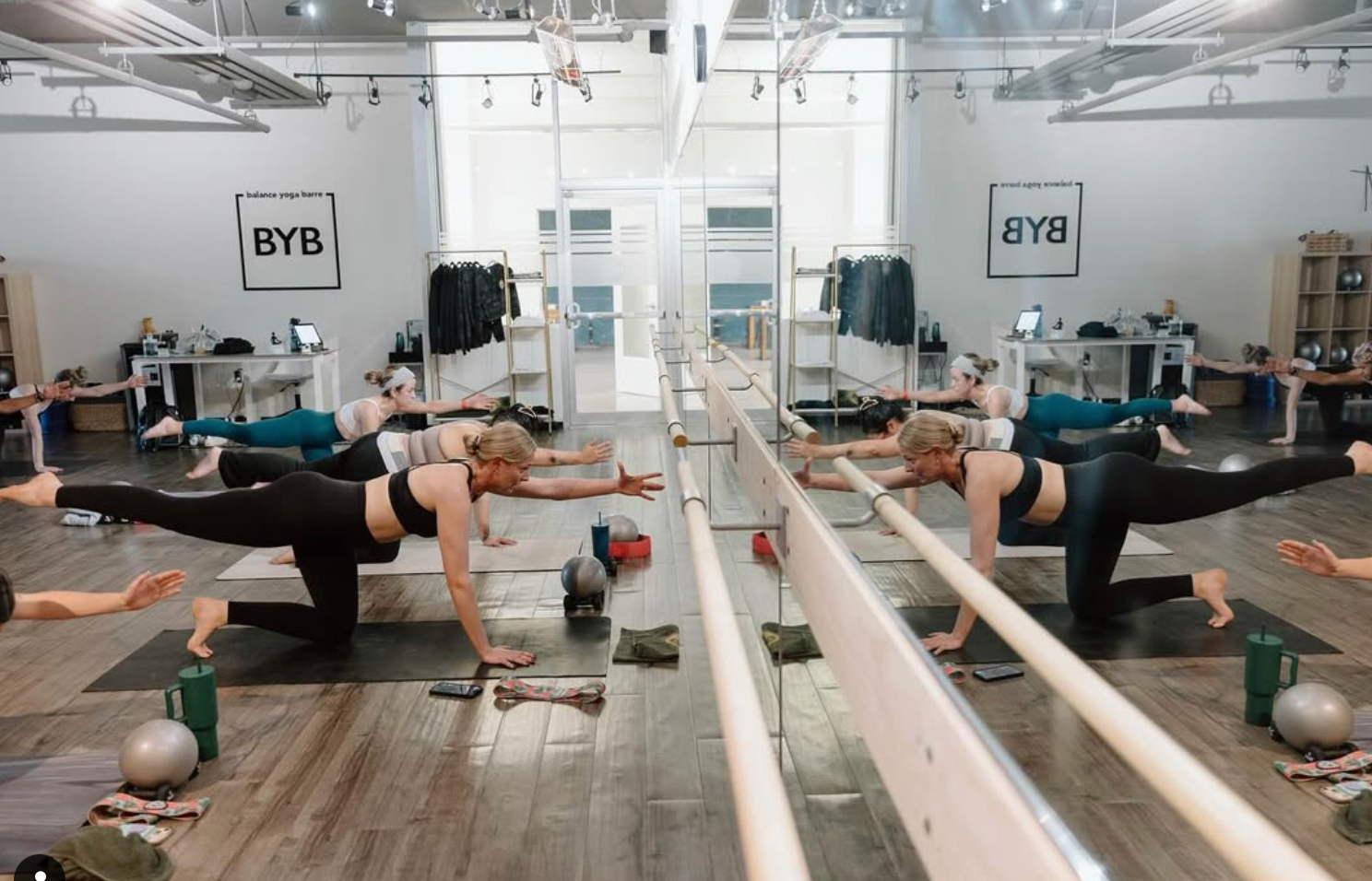 People in a barre class doing exercises, reflected in a long mirror. Wooden floor, white walls.