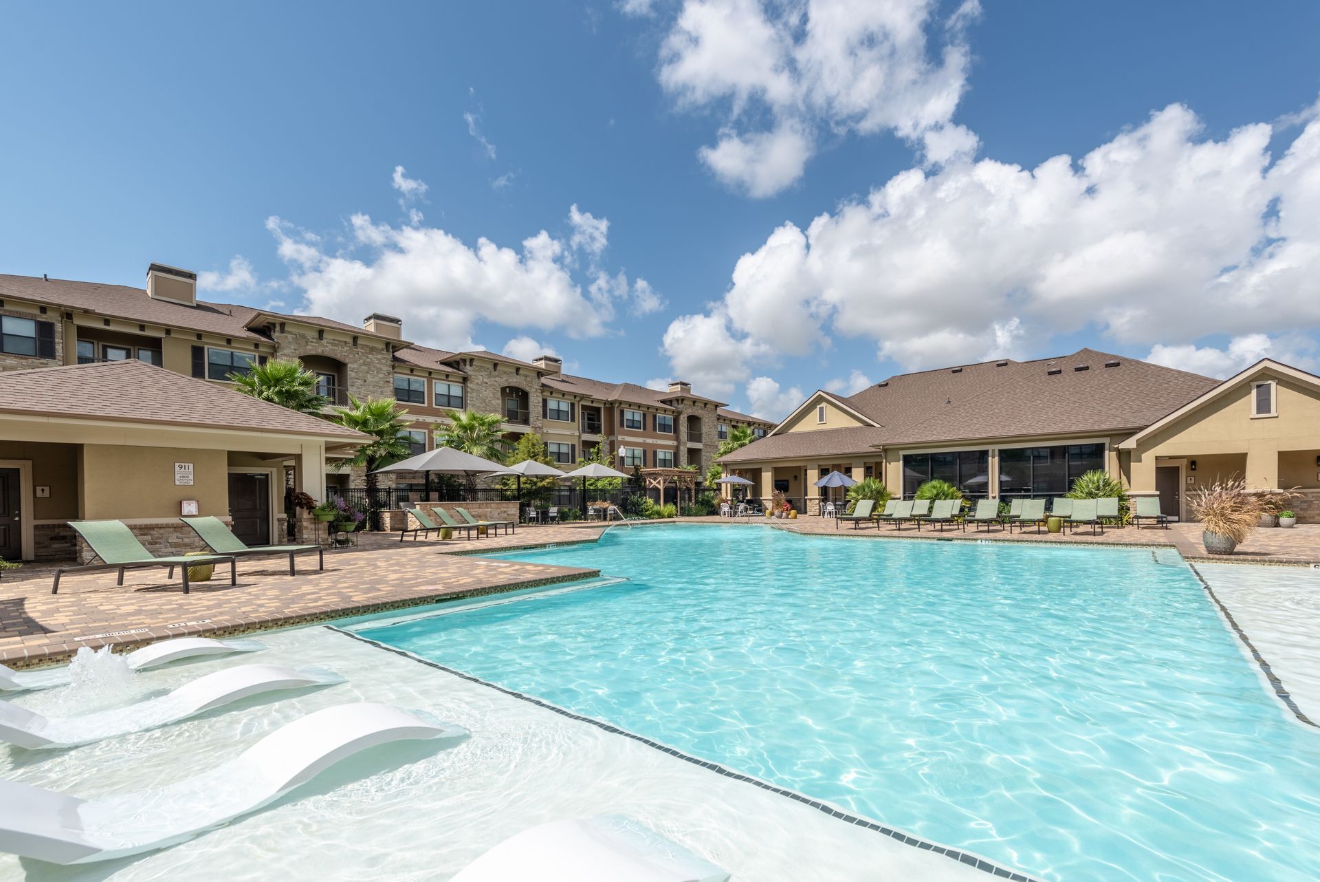 A large swimming pool surrounded by chairs and umbrellas in front of a building at Marquis Seven Lakes offers apartments for rent near Cinco Ranch, TX.
