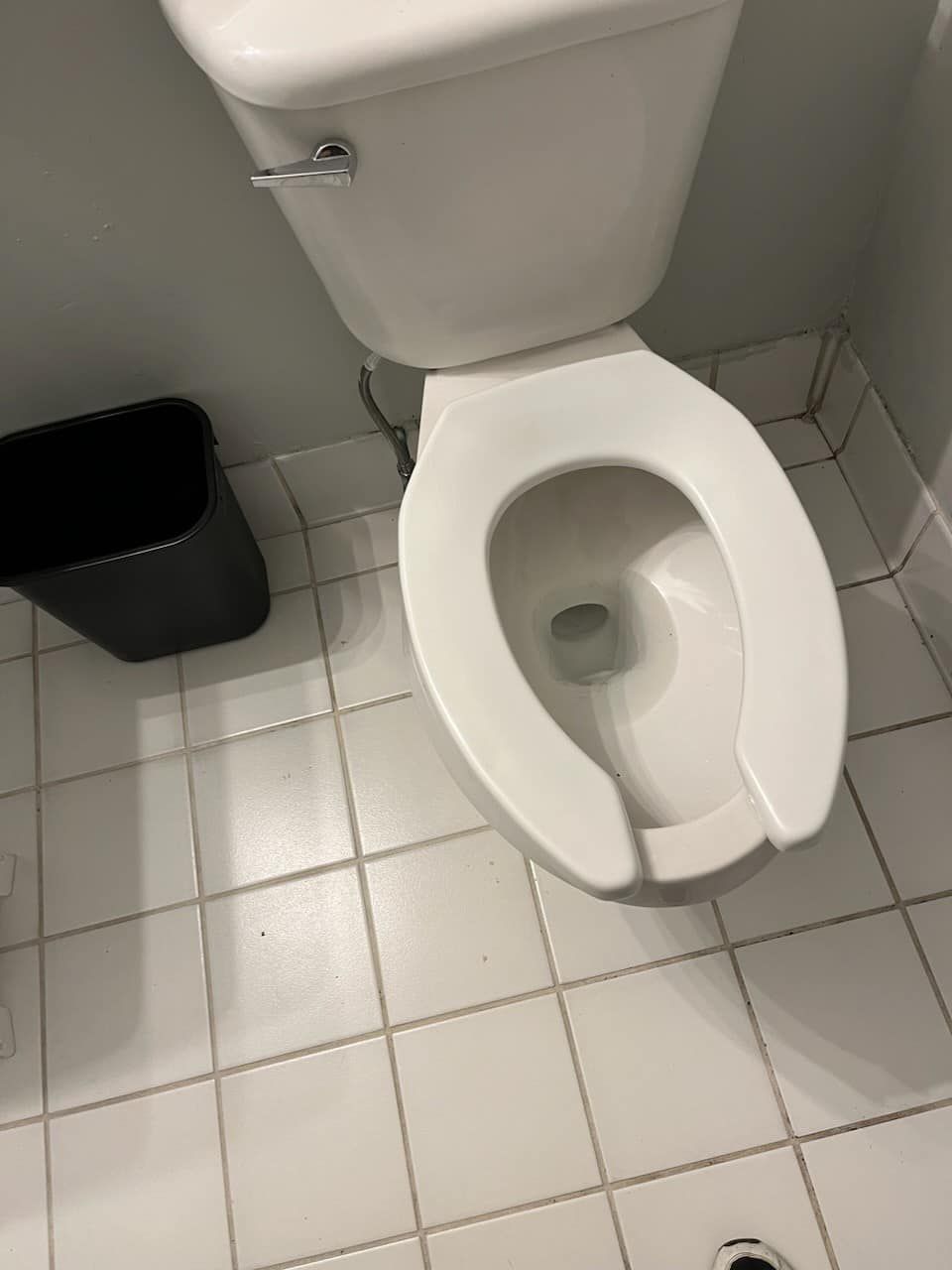 White toilet with open seat, set against white tiled floor, black trash can next to it.