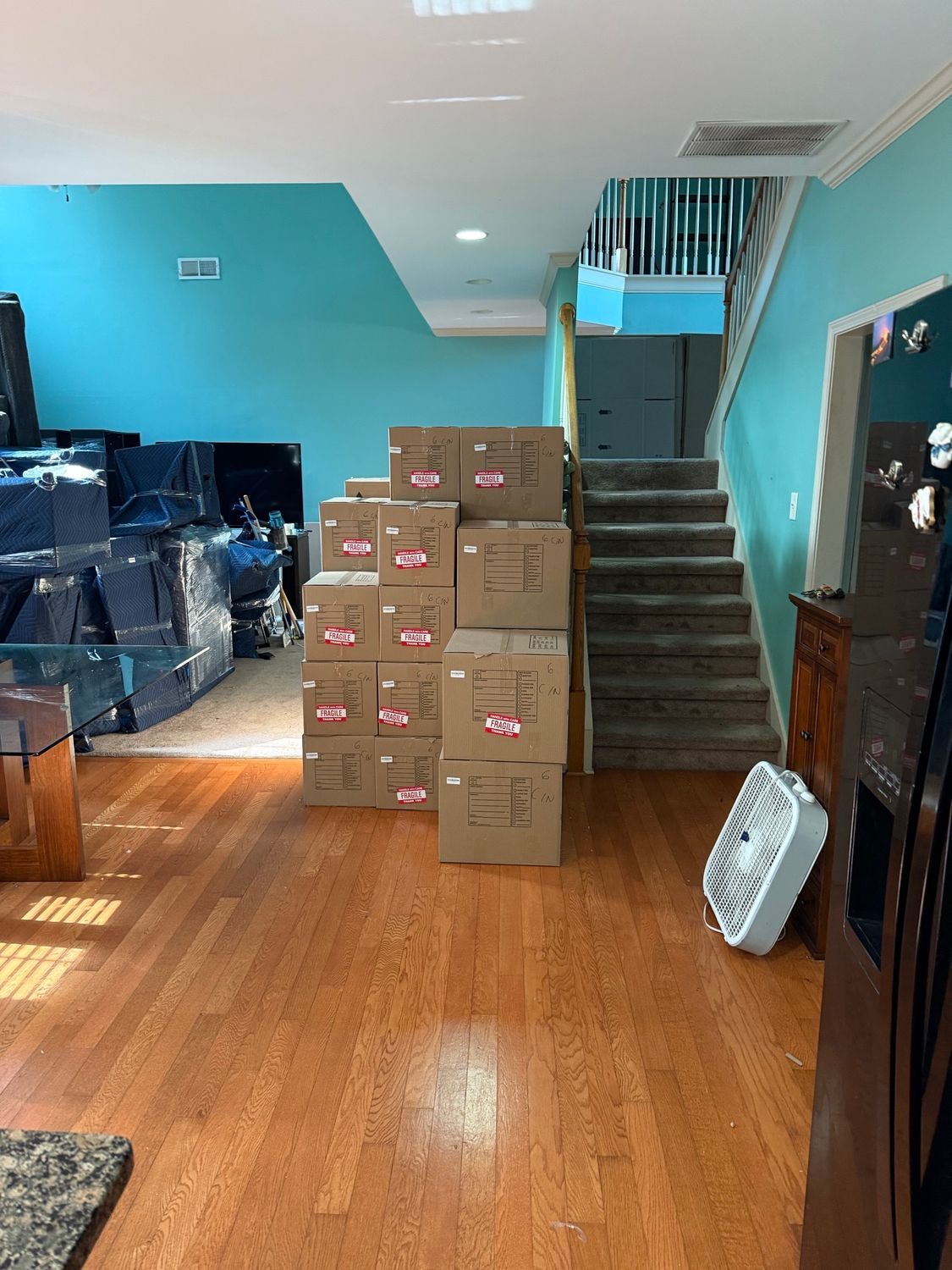 Boxes stacked in a house with wood floors, blue walls, and stairs. Furniture wrapped in plastic is in the background.