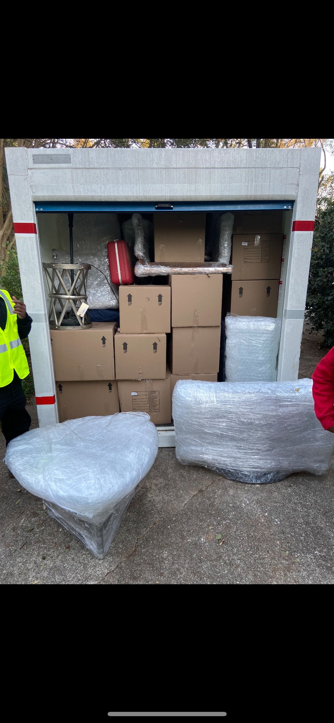 Boxes and furniture in bubble wrap inside a storage unit. Someone in a safety vest is nearby.