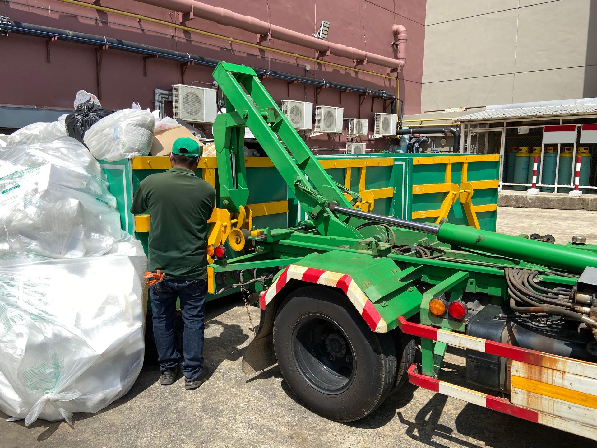 Man loading white bags into a green skip truck outside a building.