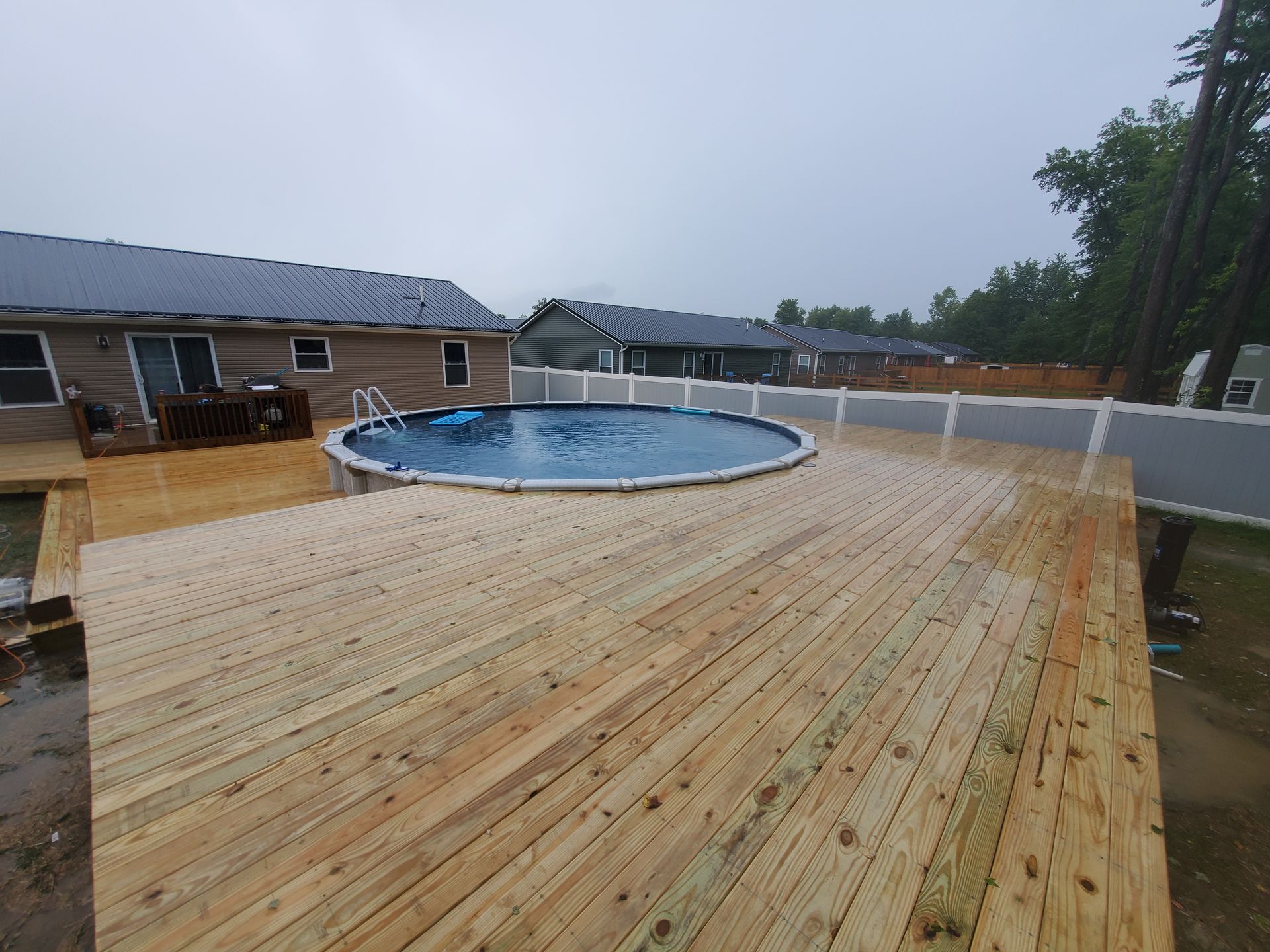 A backyard with a wooden deck surrounding a circular above-ground pool. Houses and a fence are visible.