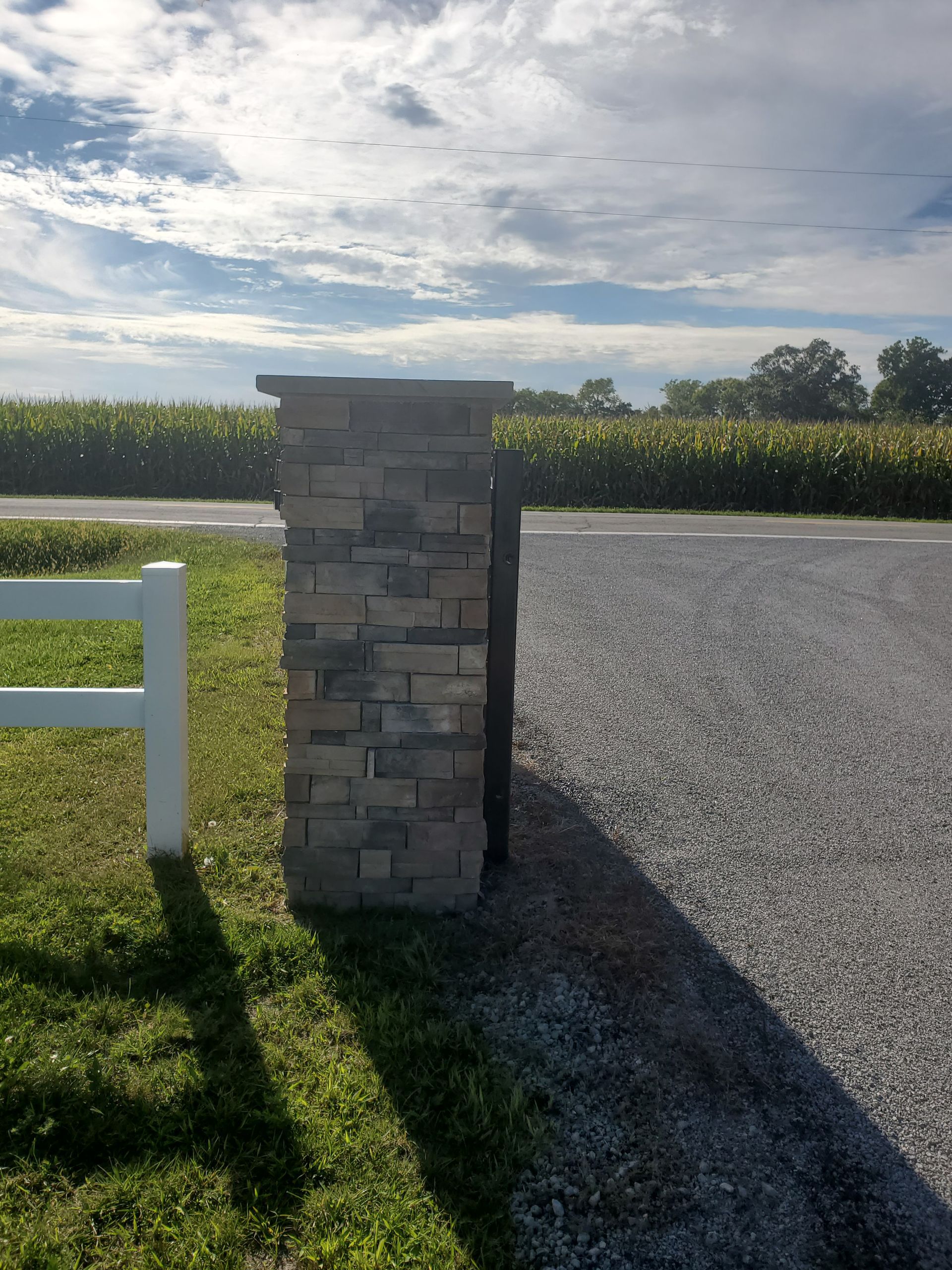Stone column next to a white fence, on a gravel drive with a green field and sky in the background.