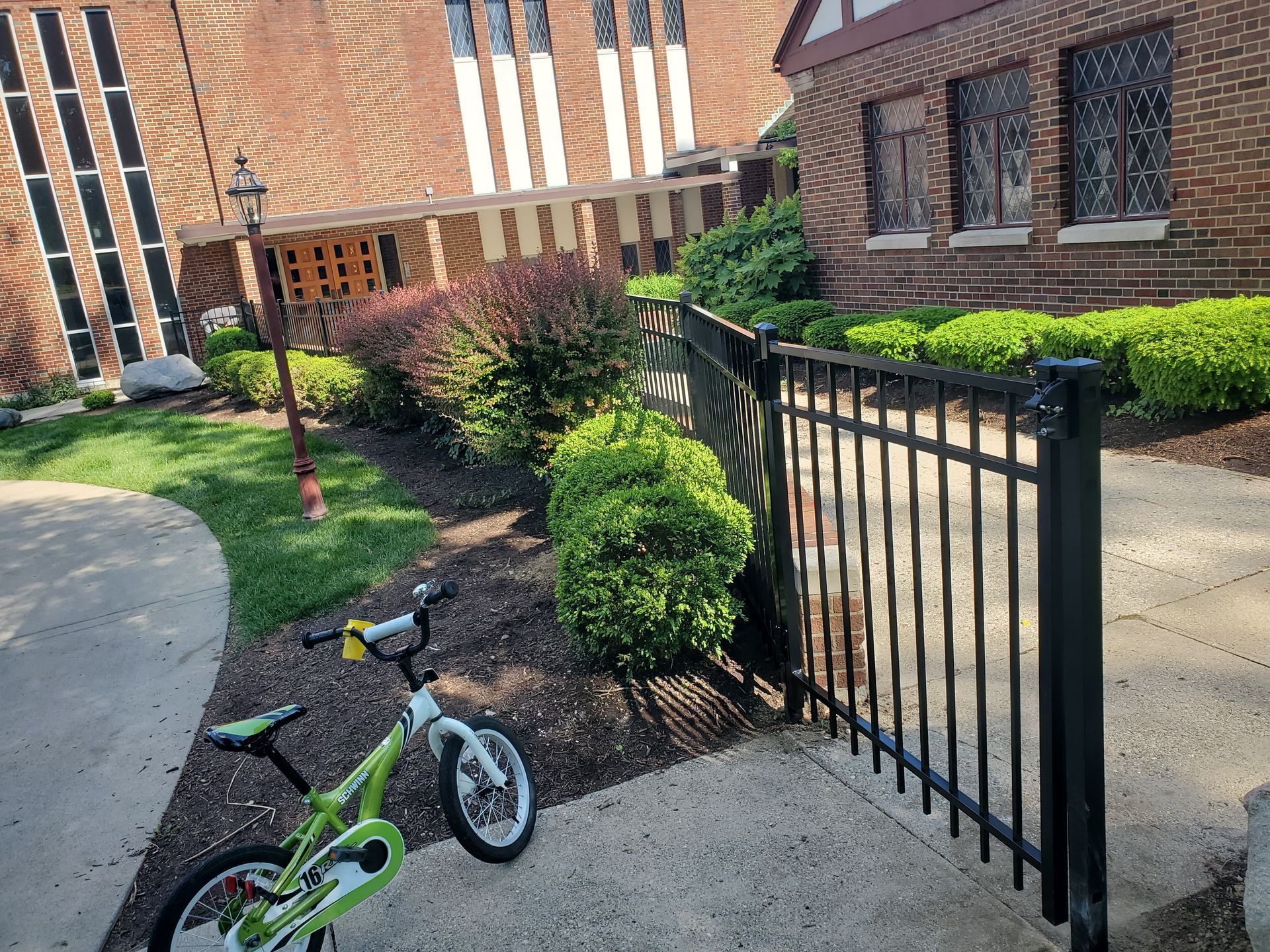 A green bicycle near a black fence, bushes, and a brick building.