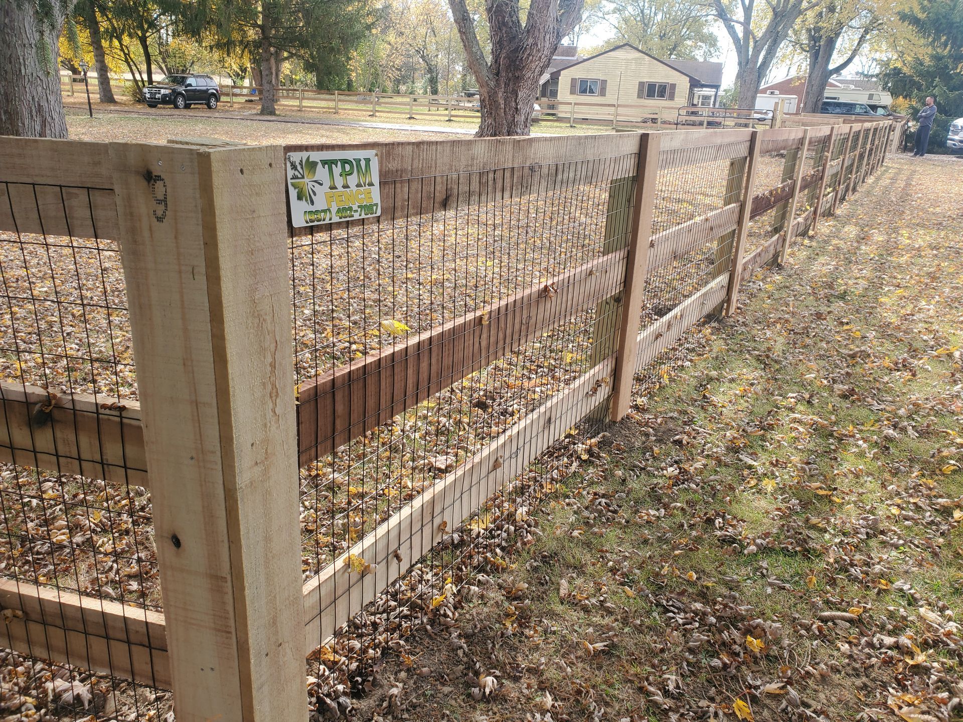 Wooden fence with wire mesh, in a grassy area with fallen leaves. A sign is attached to the fence.