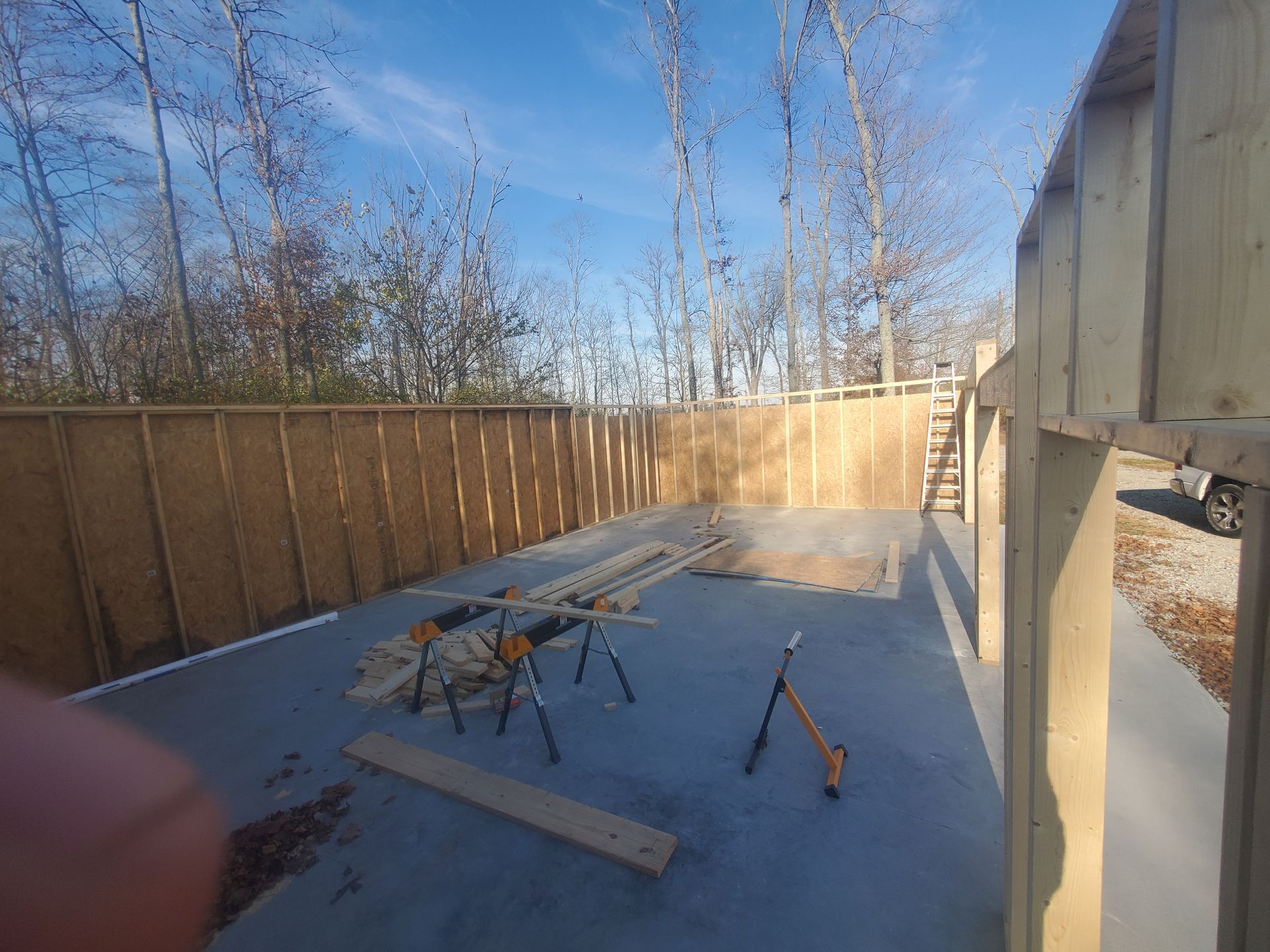 Construction site: Wooden walls and floor, tools, and lumber on a concrete base with trees in the background under a blue sky.