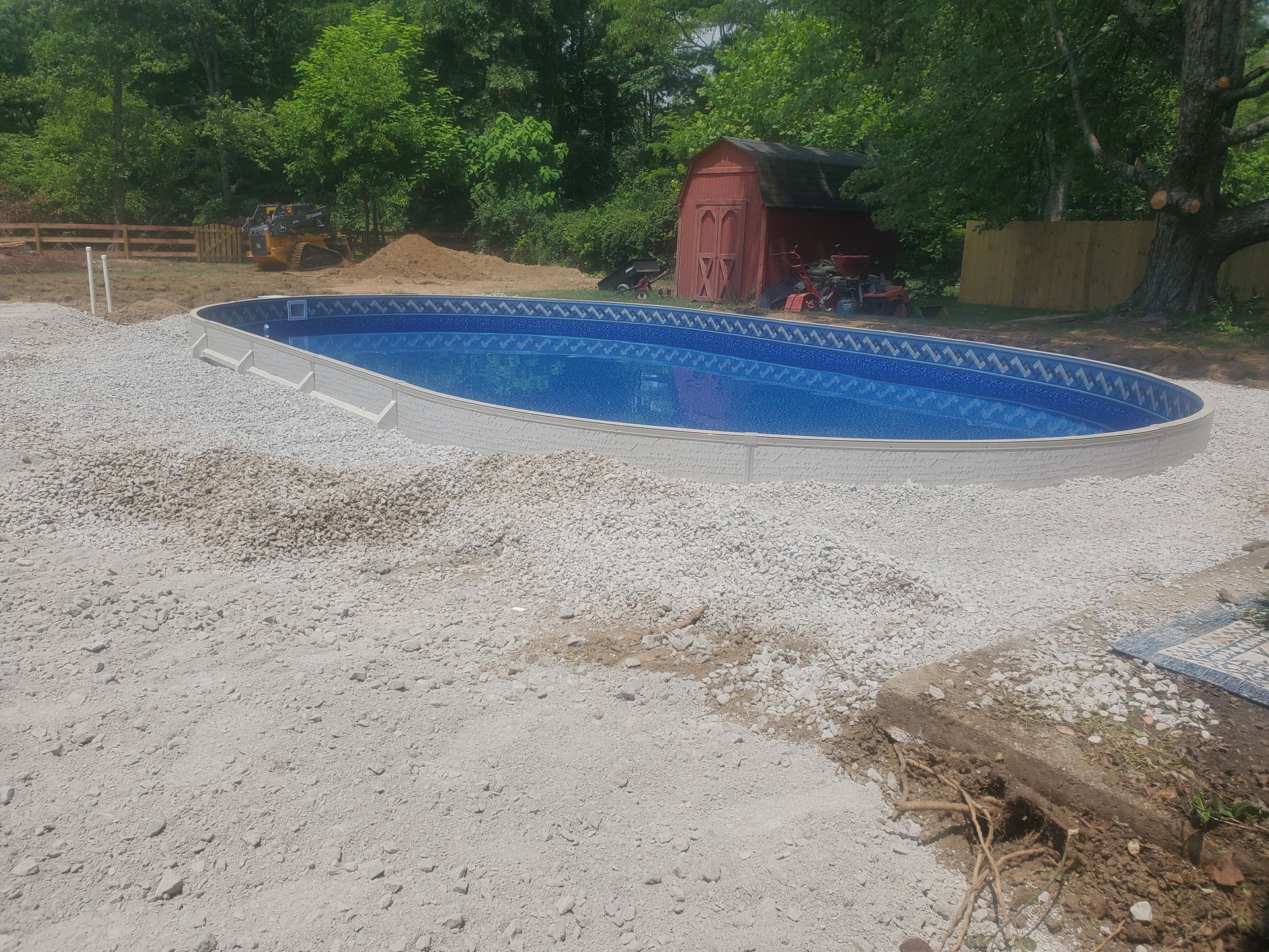 Oval-shaped, above-ground swimming pool under construction, surrounded by gravel. A small red shed is visible in the background.